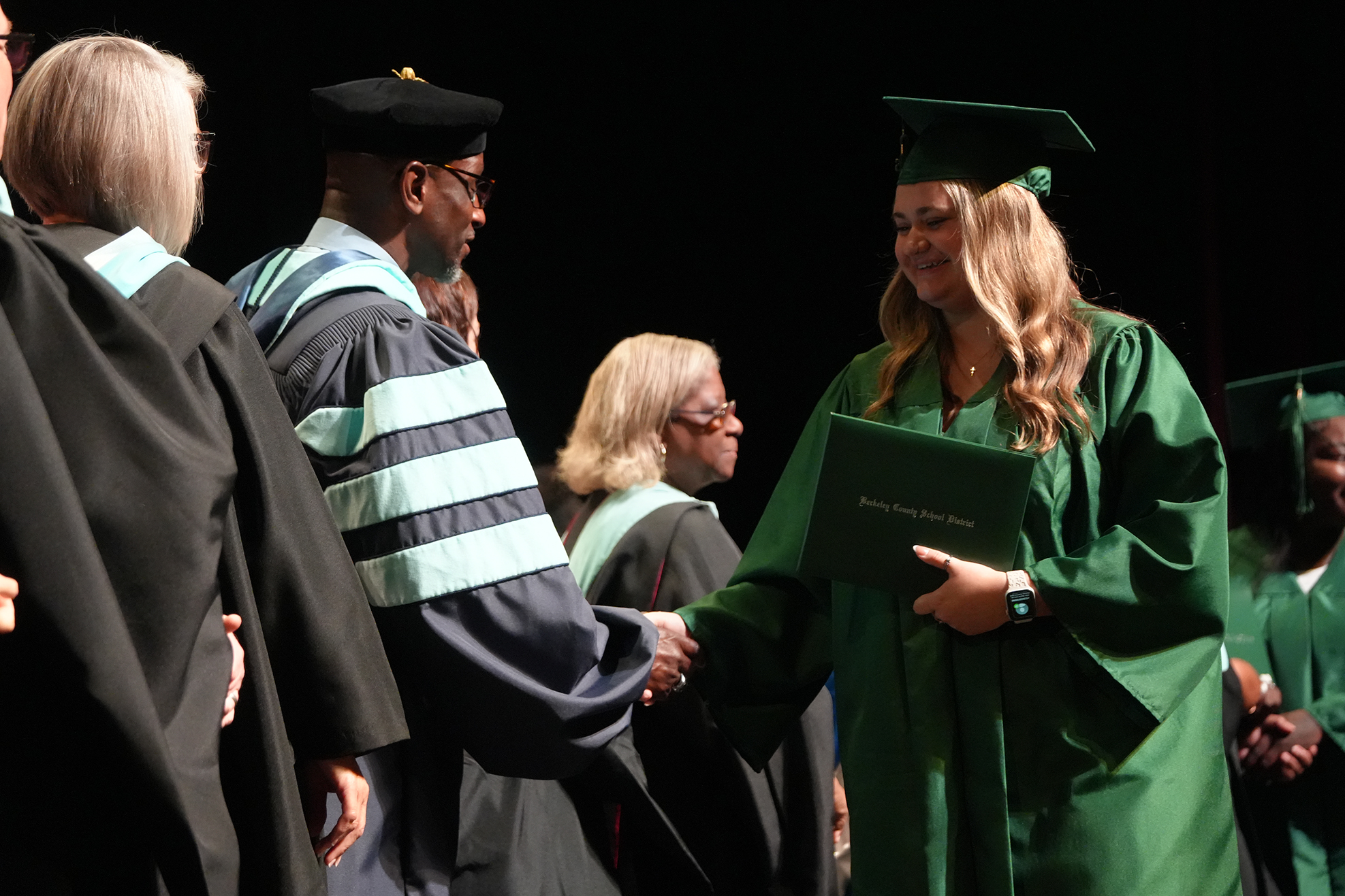 A woman in a green graduation gown shakes hands with a man in a blue and gray gown.