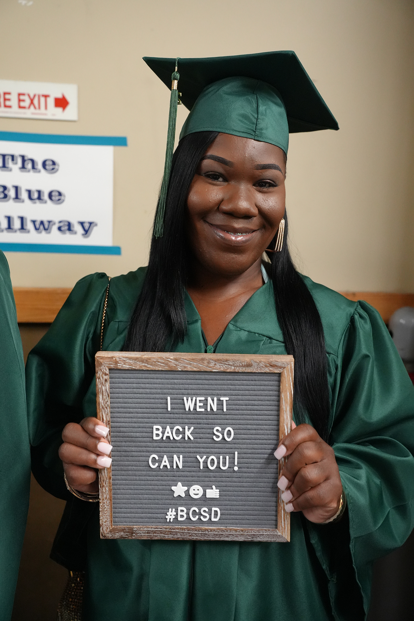 A woman in a green graduation gown holds a sign that says, "I WENT BACK SO YOU CAN TOO! #BCSD."