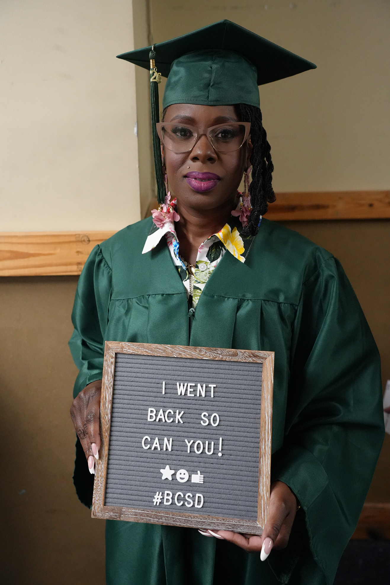 Woman in green graduation gown holding a sign that reads, "I WENT BACK SO CAN YOU! #BCSGrad"