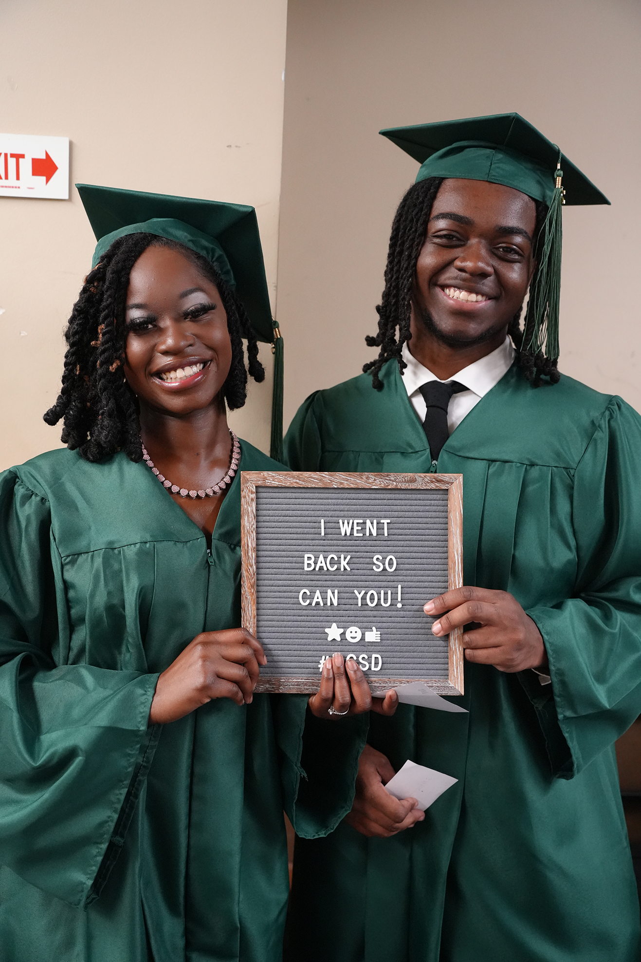 Two people in green graduation gowns hold a sign saying "I WENT BACK SO YOU CAN YOU!" with smiles.