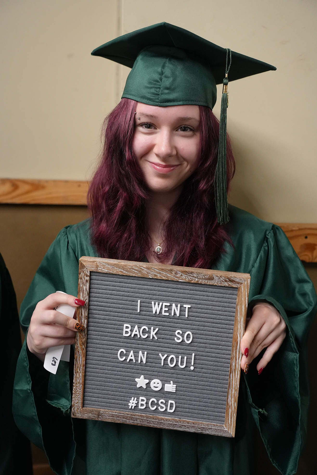 A person in a green gown and cap holds a chalkboard sign with white text.