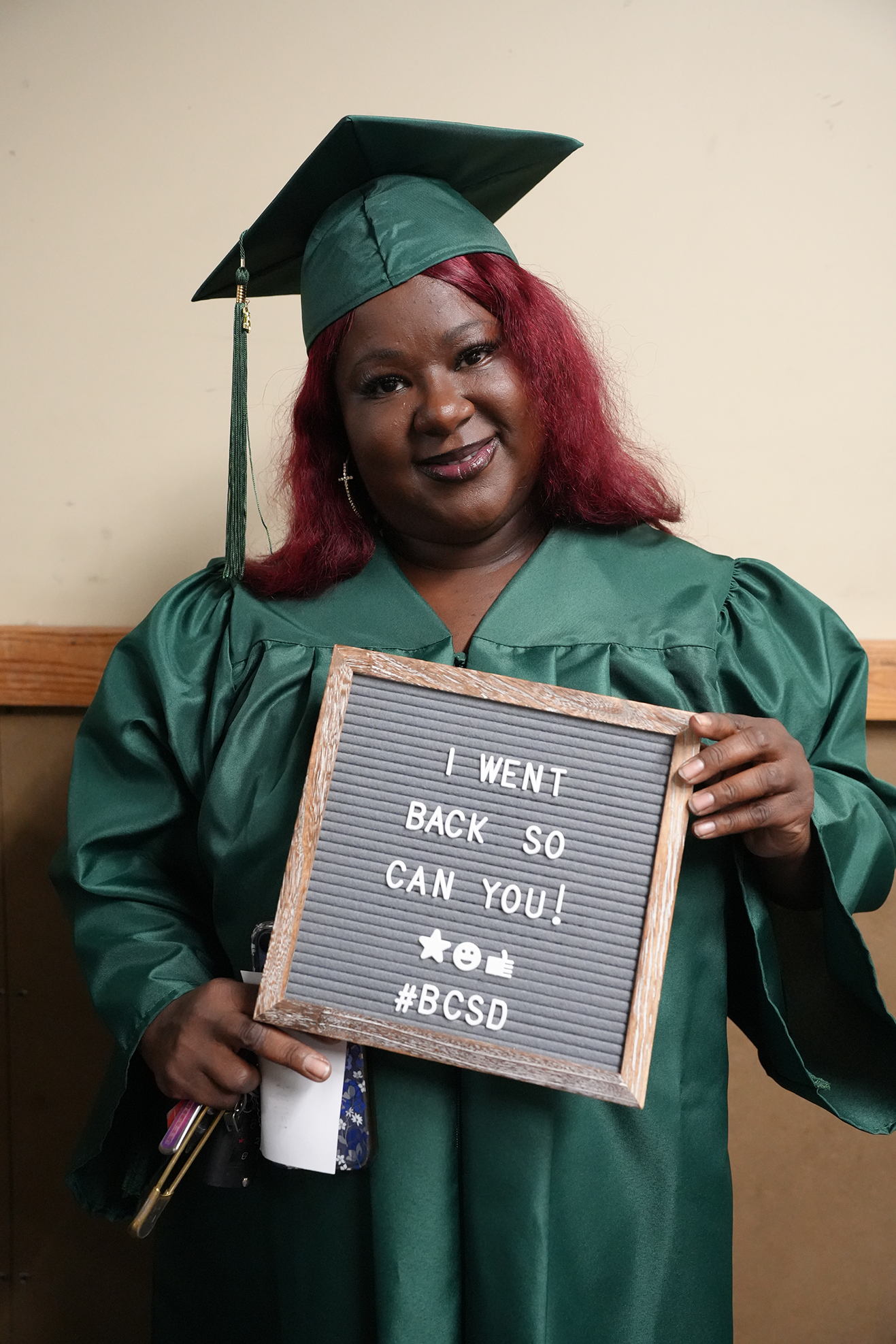 Person in green graduation gown and cap, holding a sign reading "I WENT BACK SO CAN YOU! #BCSDU".
