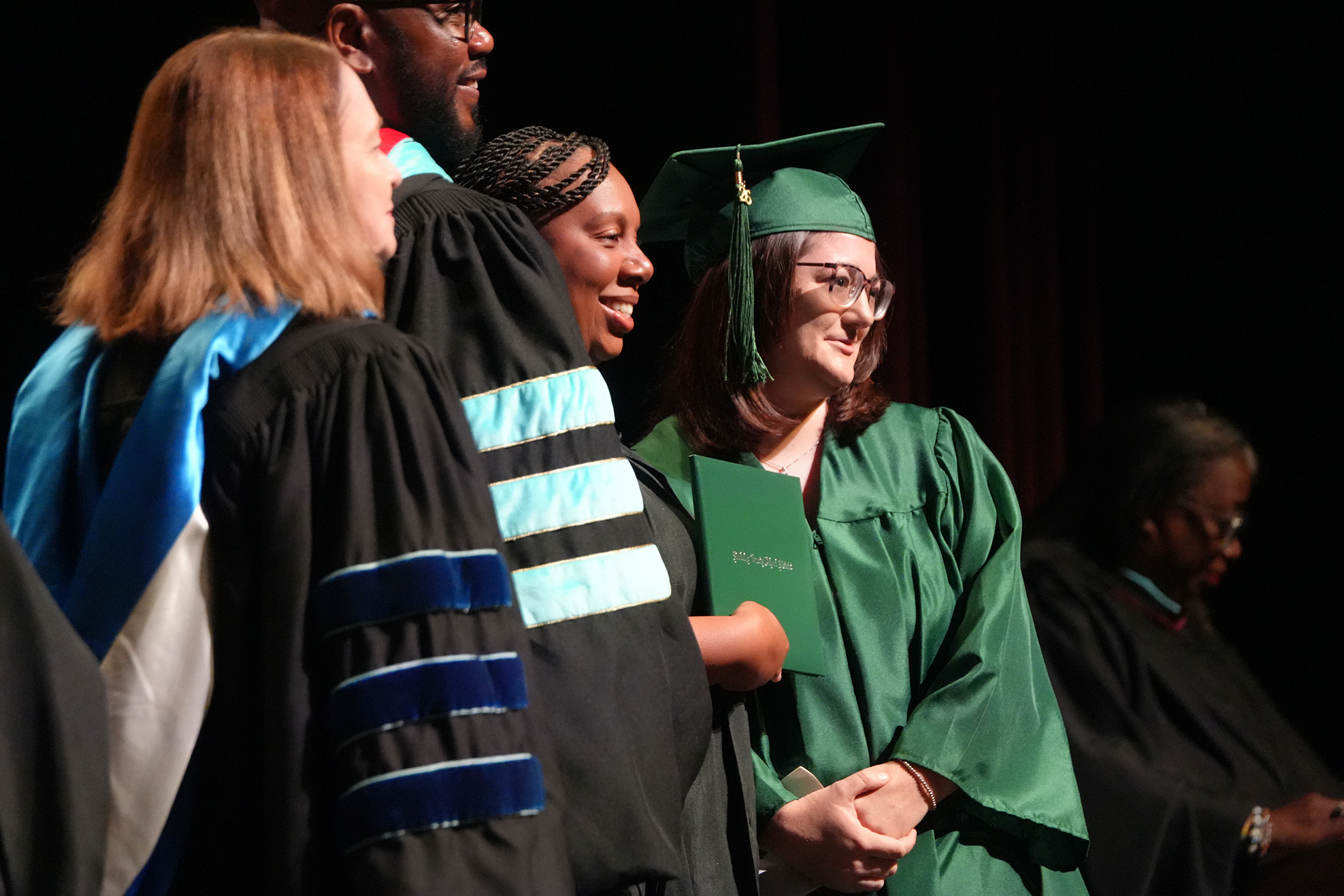 A group of people in caps and gowns on stage, one woman smiling, others talking. A blurred figure with glasses in the background.