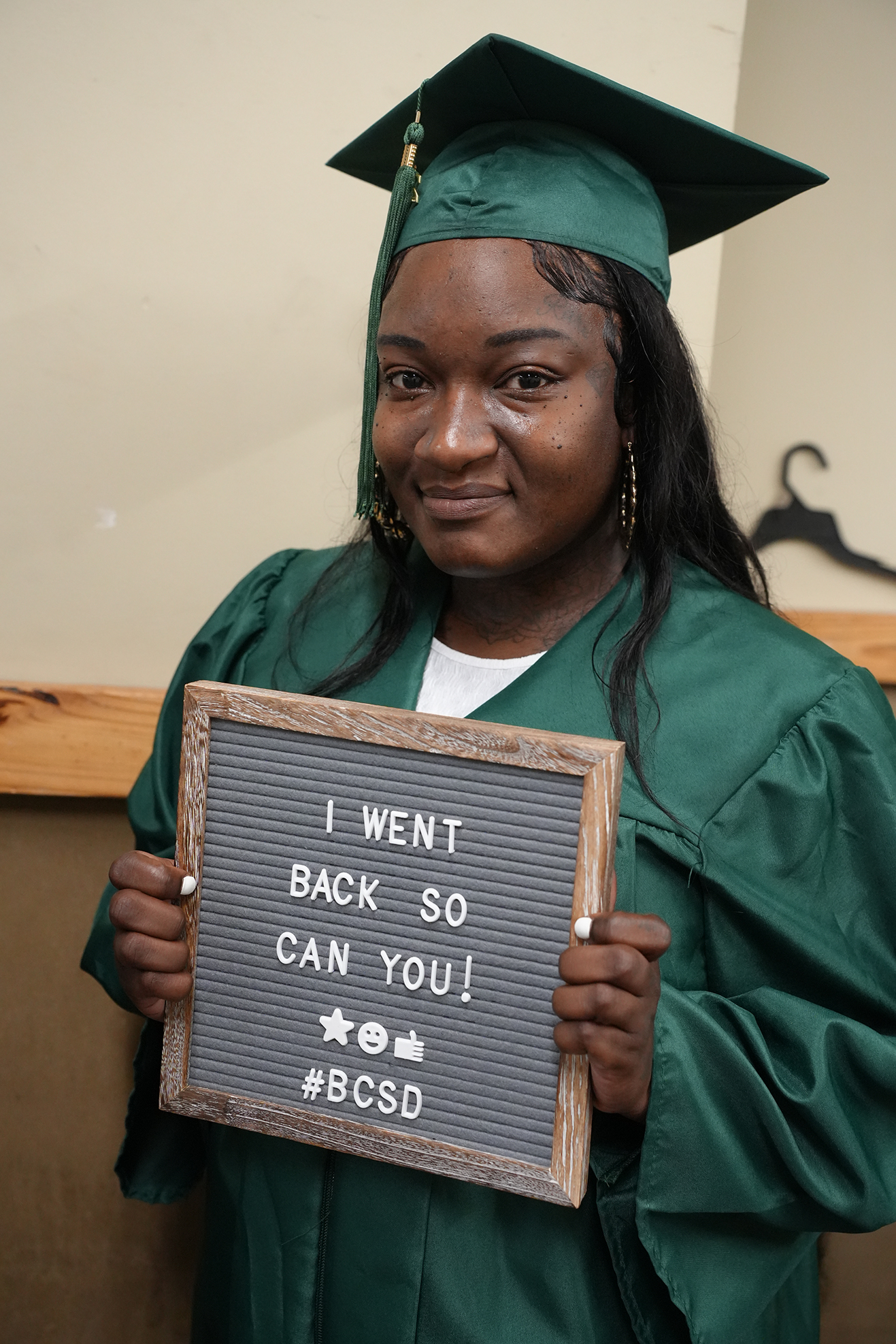 A woman in a green graduation gown holds a chalkboard sign saying "I WENT BACK SO CAN YOU! #BCSd."