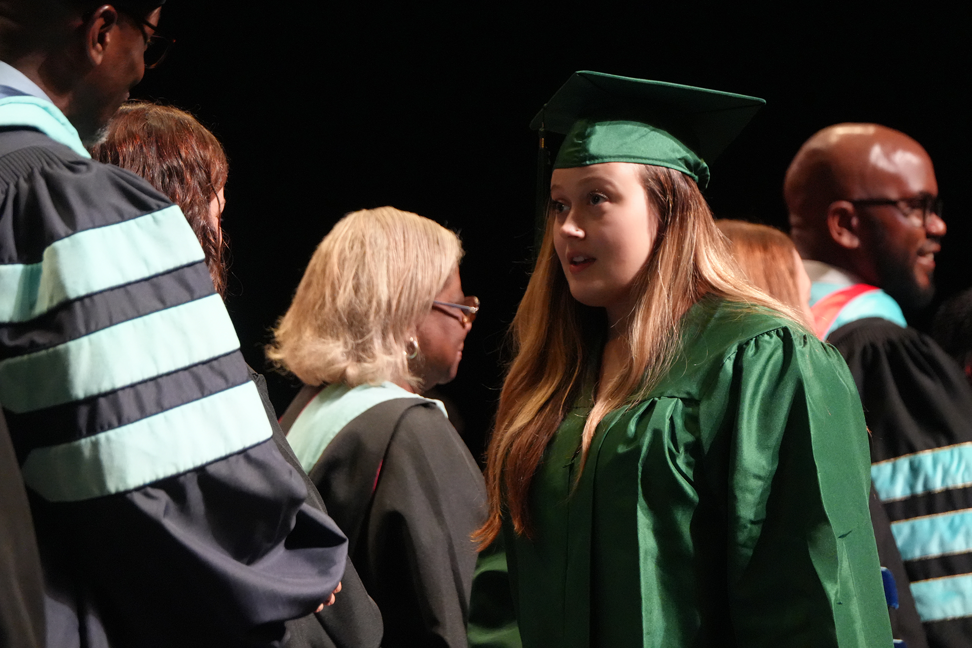 A woman in a green graduation gown stands among others in academic regalia, facing forward, likely at a commencement ceremony.