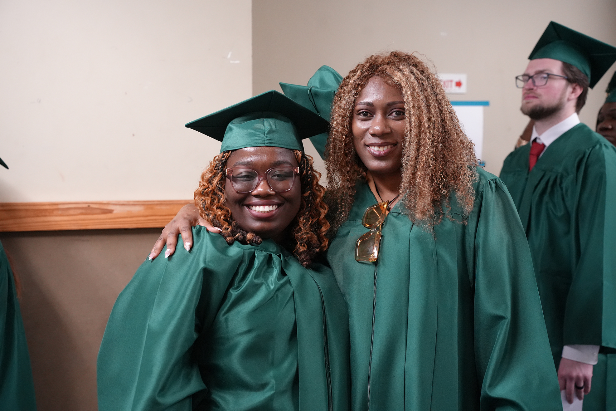 Two women in green caps and gowns pose for a photo. Behind them, a man in a similar outfit stands.