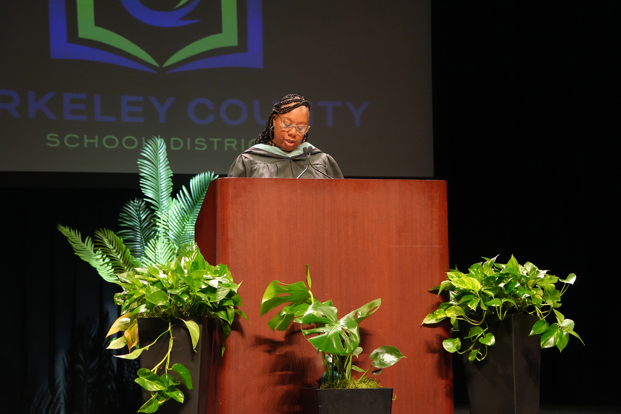 A person stands at a podium with green plants, speaking. A Berkeley County School District logo is displayed behind.