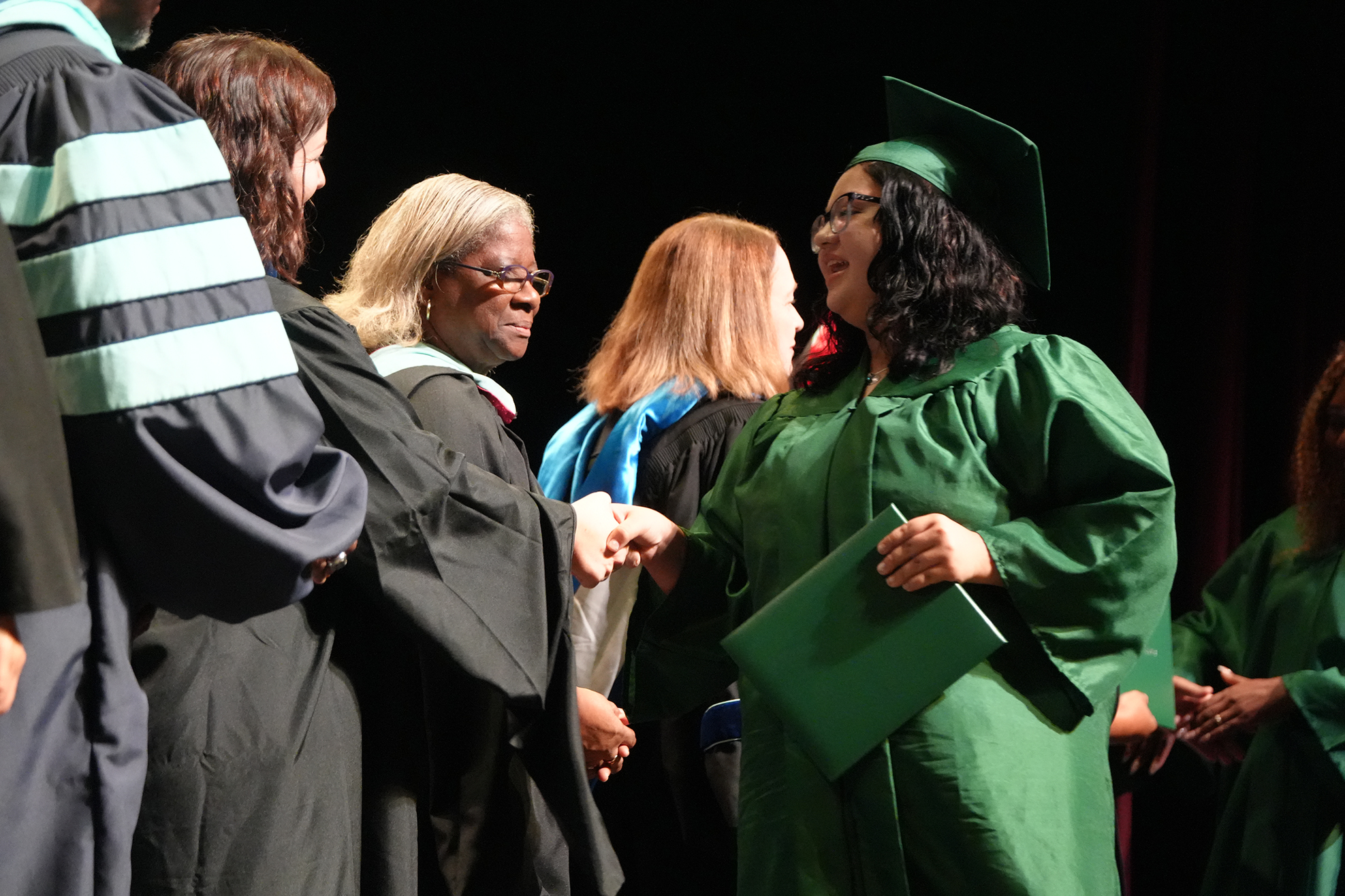 Graduates in green gowns and caps on a stage, one holding a folder, others in black gowns.