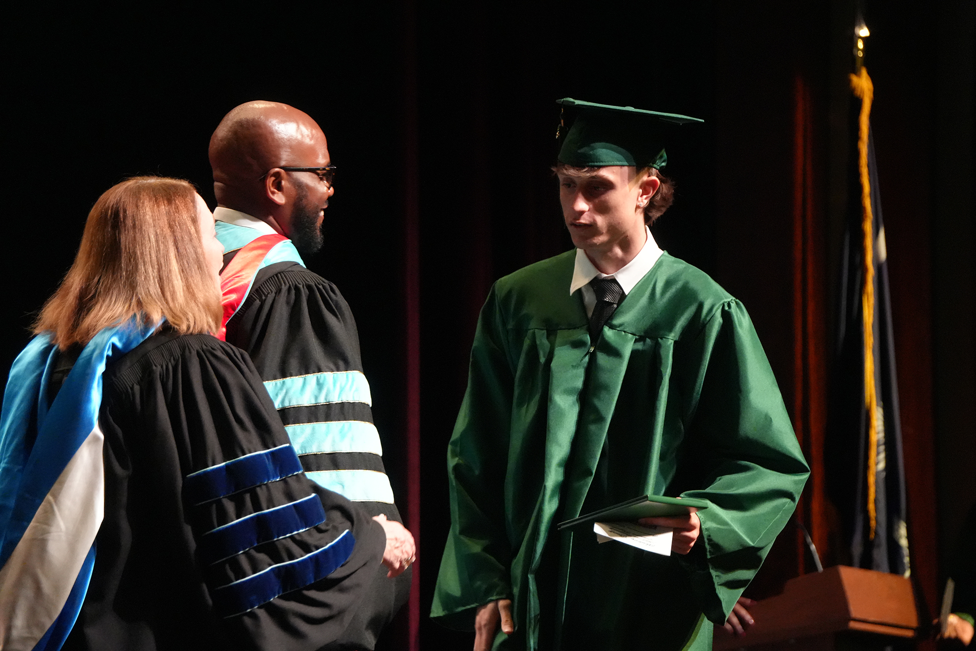 A man in a green graduation gown and cap stands with a diploma, facing two others in academic regalia.