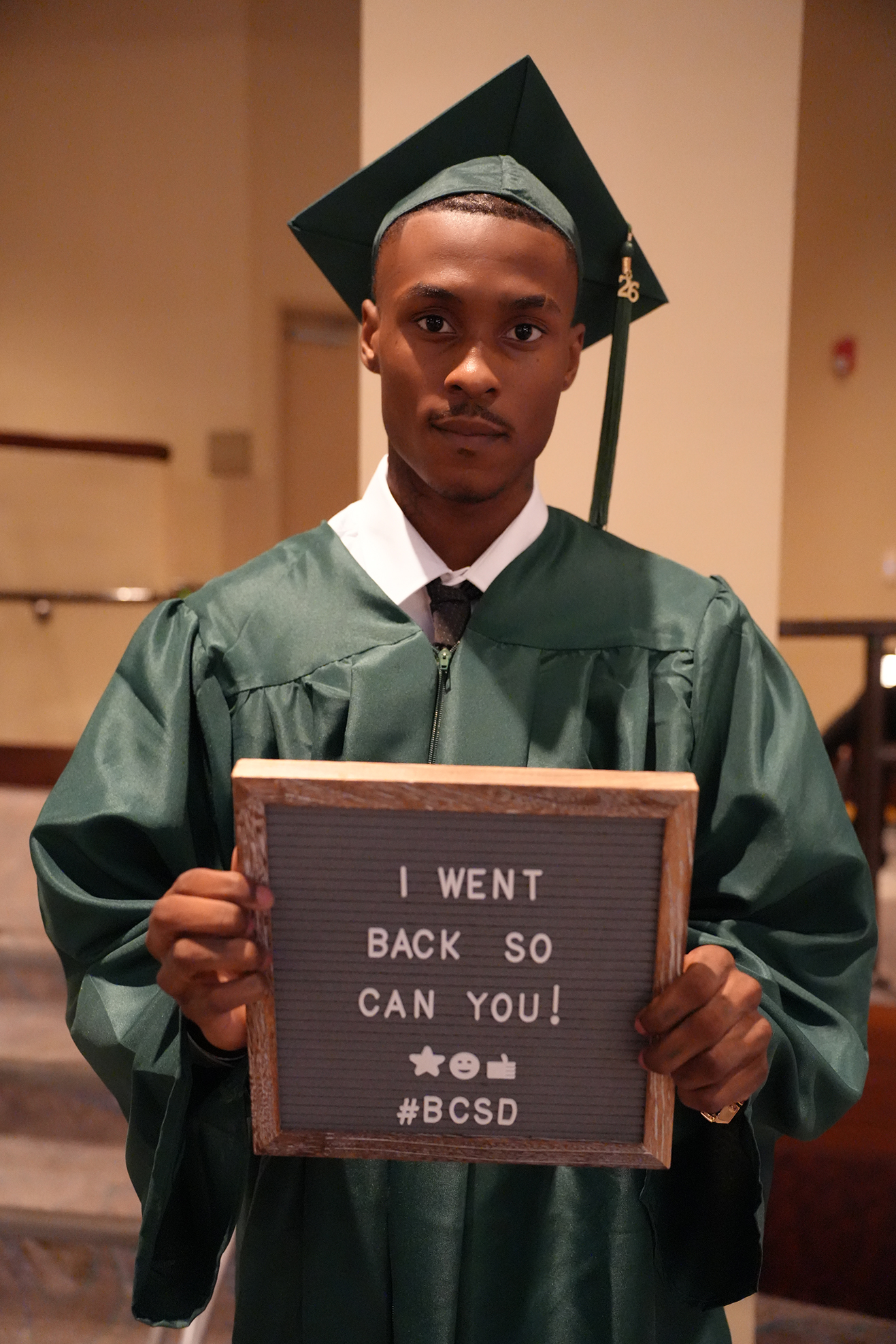 A male graduate in green gown and cap holds a sign that reads "I WENT BACK SO I CAN YOU!" indoors.