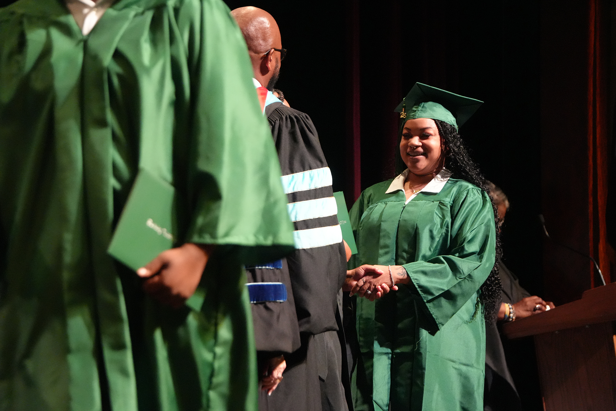A woman in a green graduation gown stands on stage, shaking hands with a graduate in a black gown.