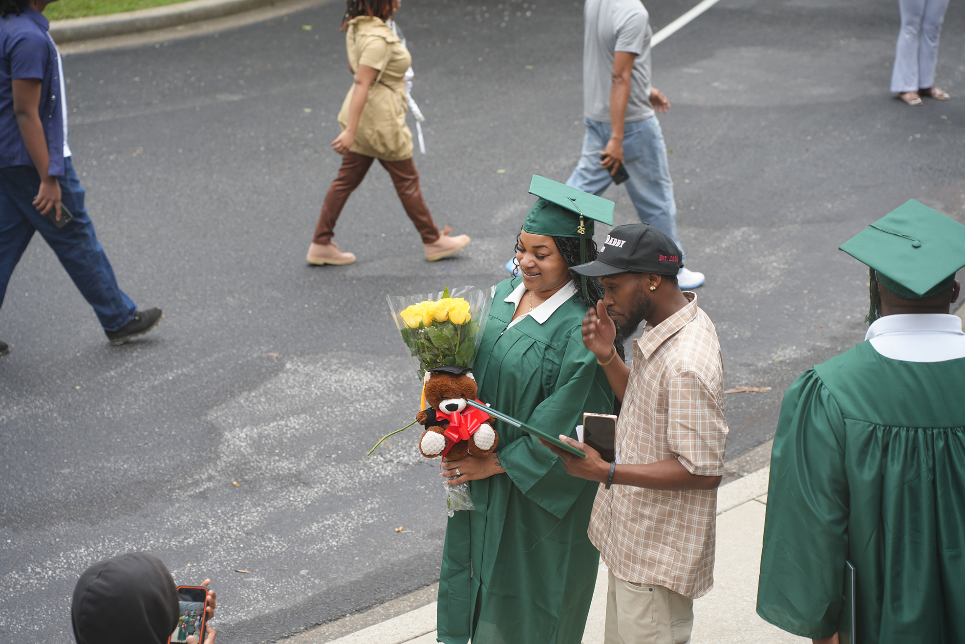 People in green graduation gowns walk on a road with one holding flowers and a stuffed animal.