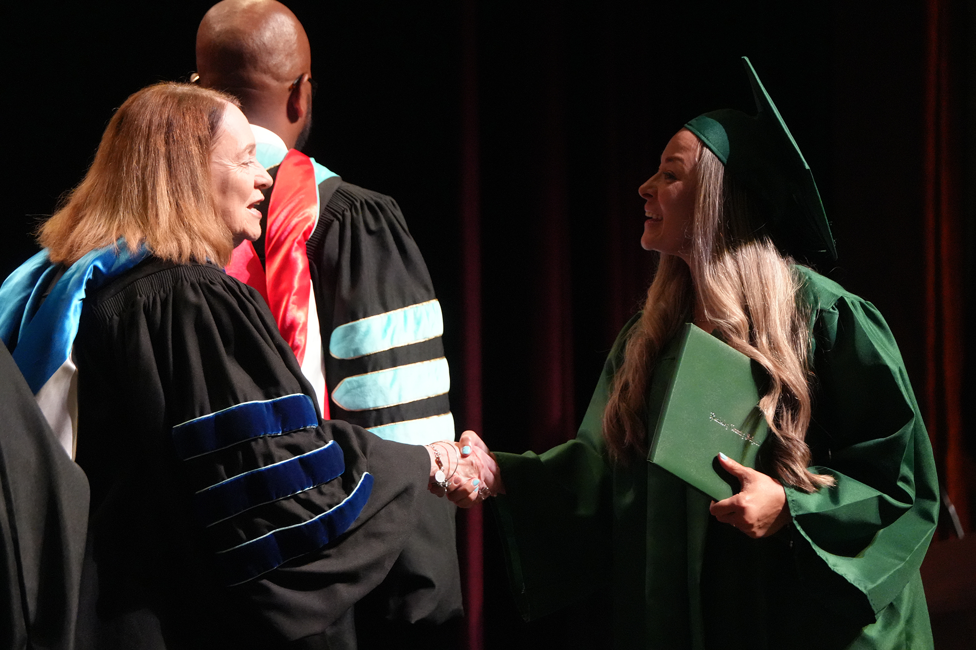 Woman in a green graduation gown shakes hands with a faculty member in a black gown.