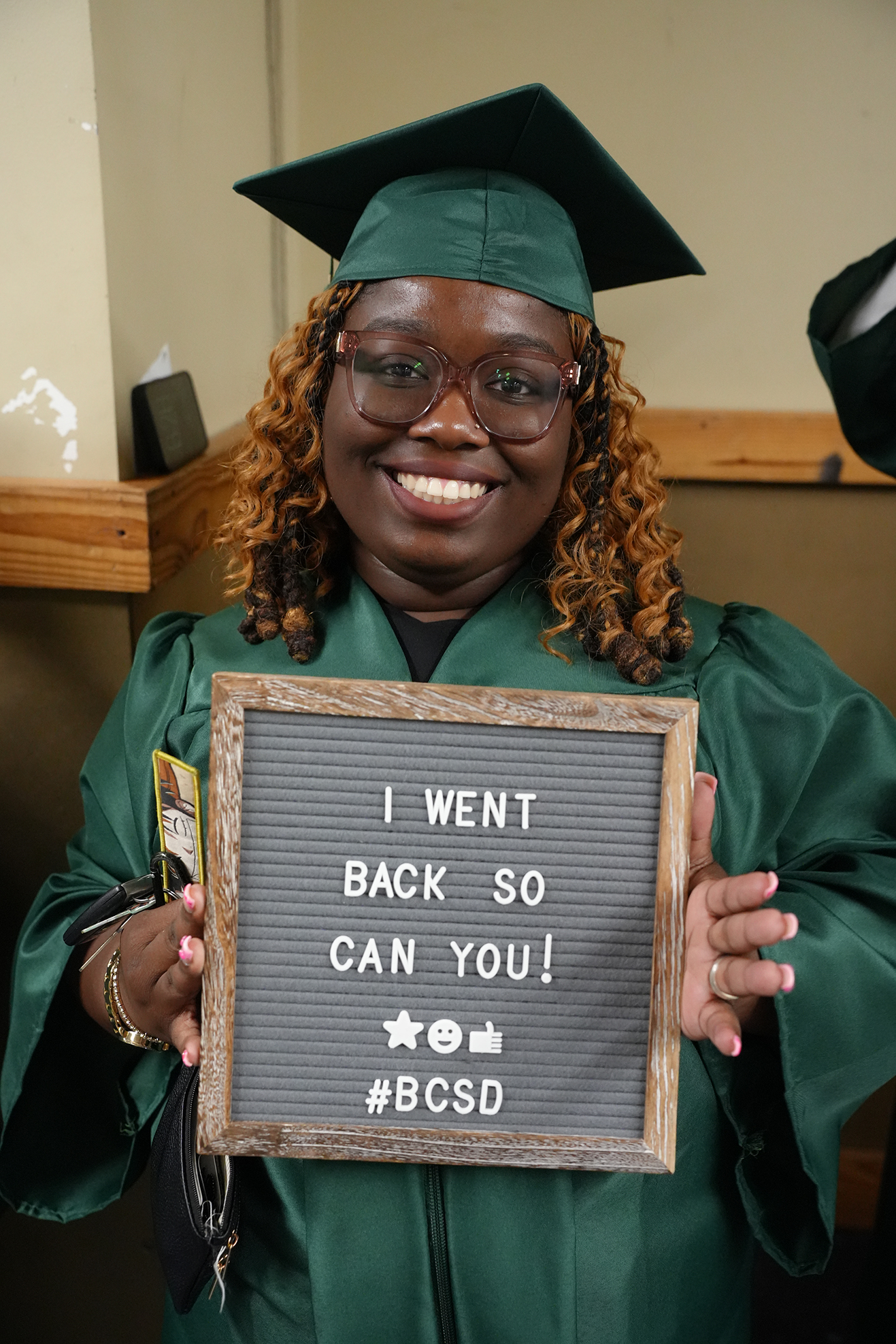 Woman in a green graduation gown holds a sign that reads "I WENT BACK SO CAN YOU!" with a smiling face.
