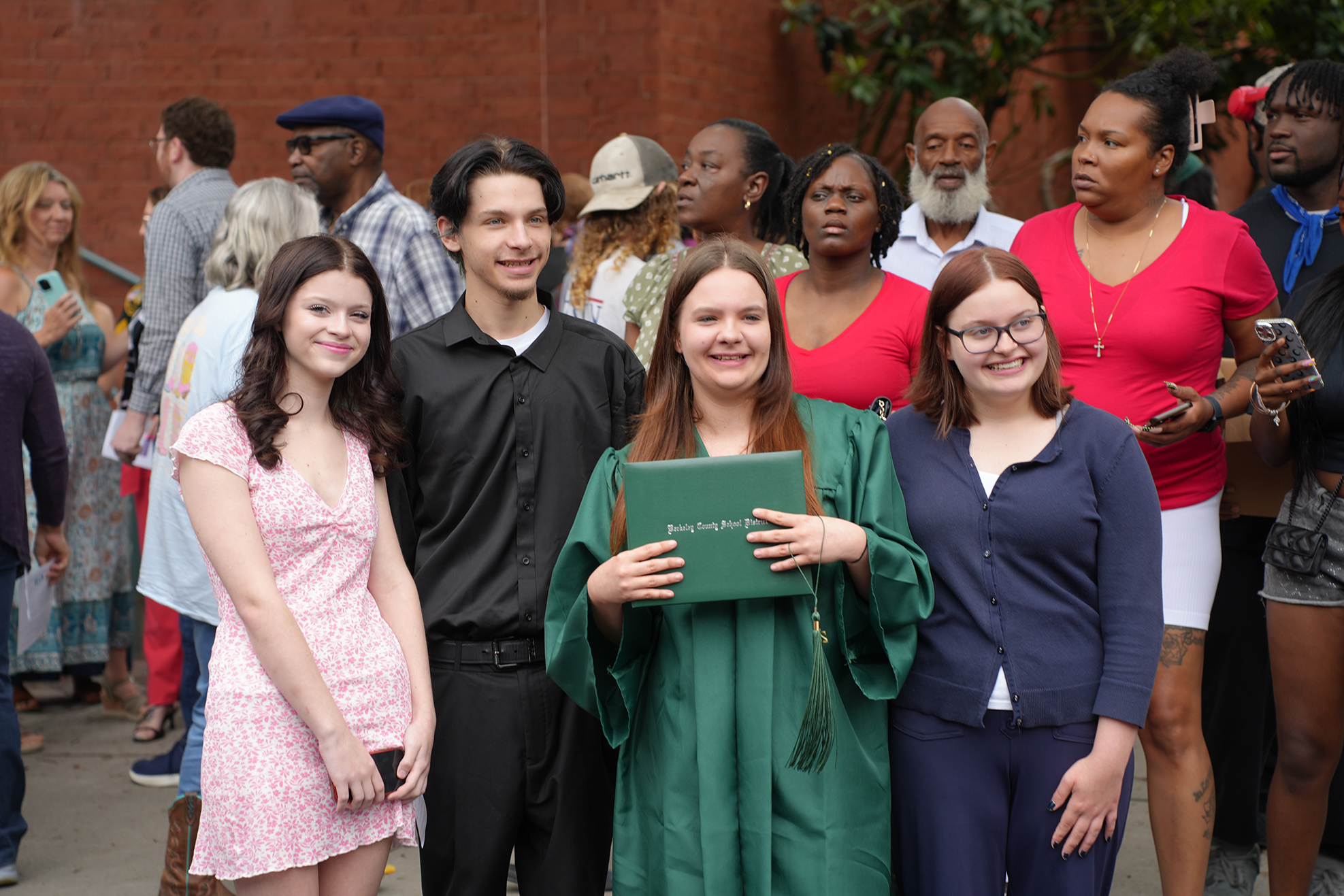 A group of people stand outdoors, some in graduation attire. A woman holds a green diploma.