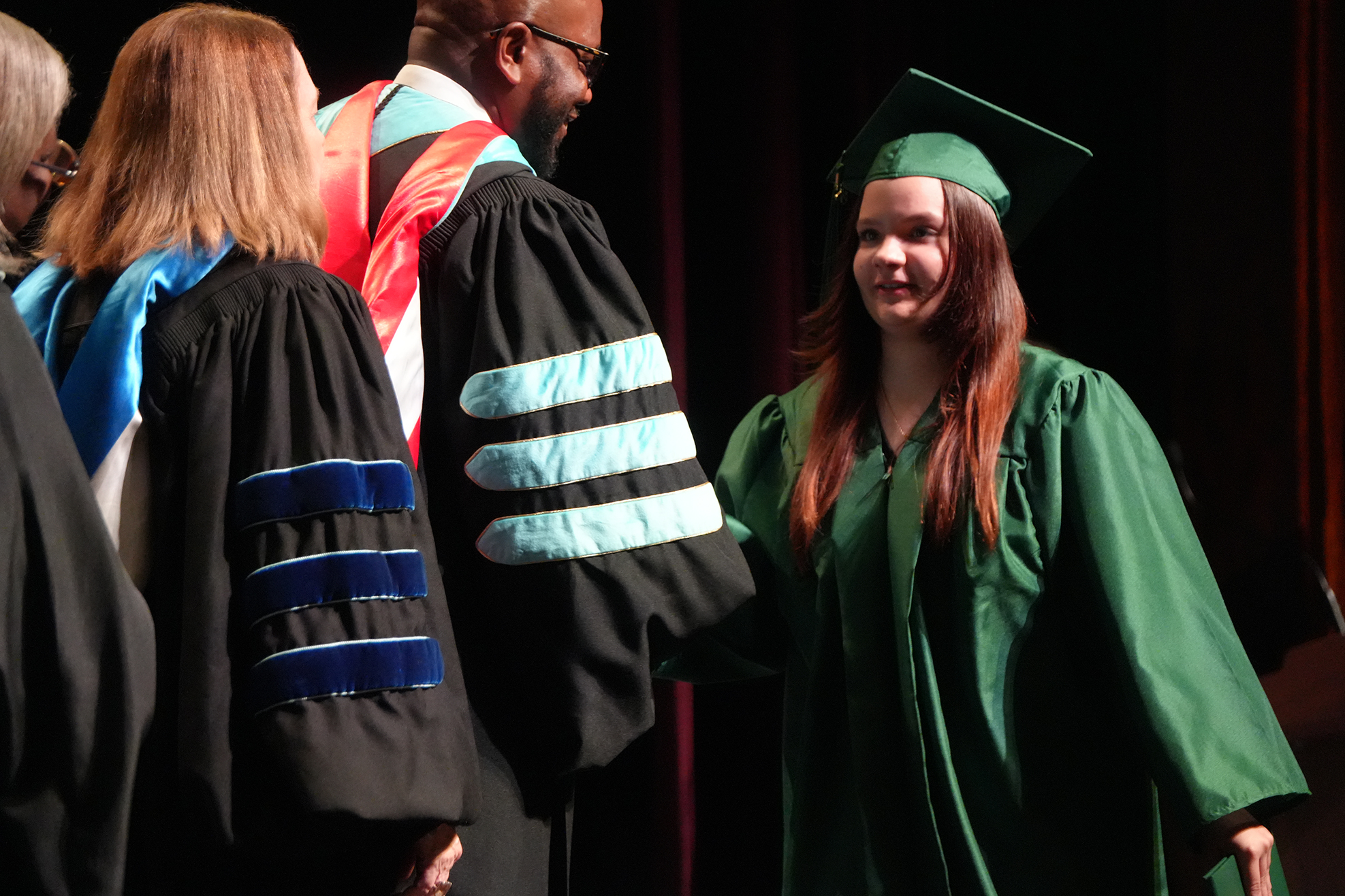 A woman in a green graduation gown smiles next to a man and woman in black gowns with blue and white trim.