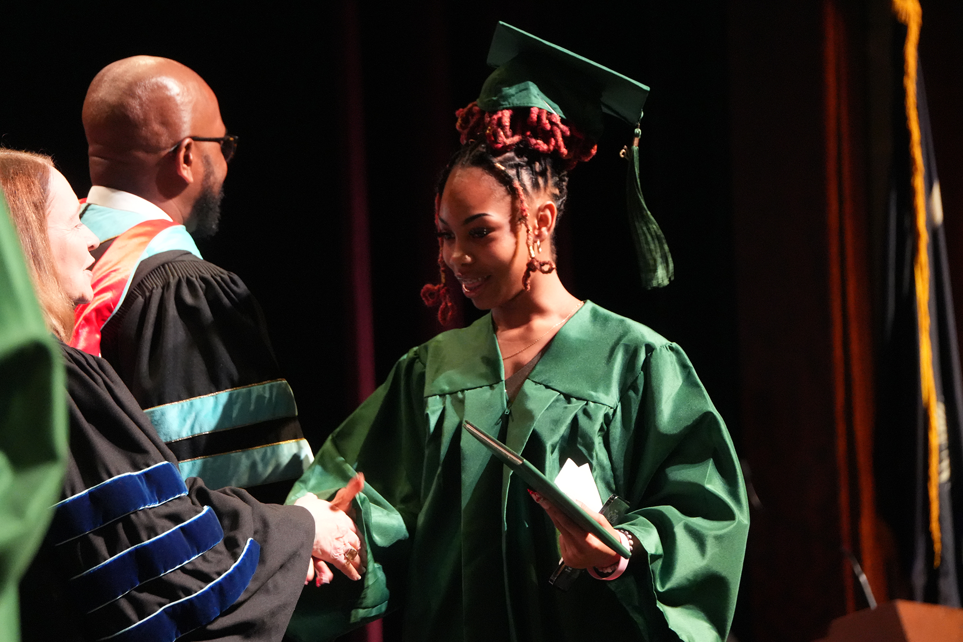 A person in a green gown and cap smiles while shaking hands with two people in black gowns.