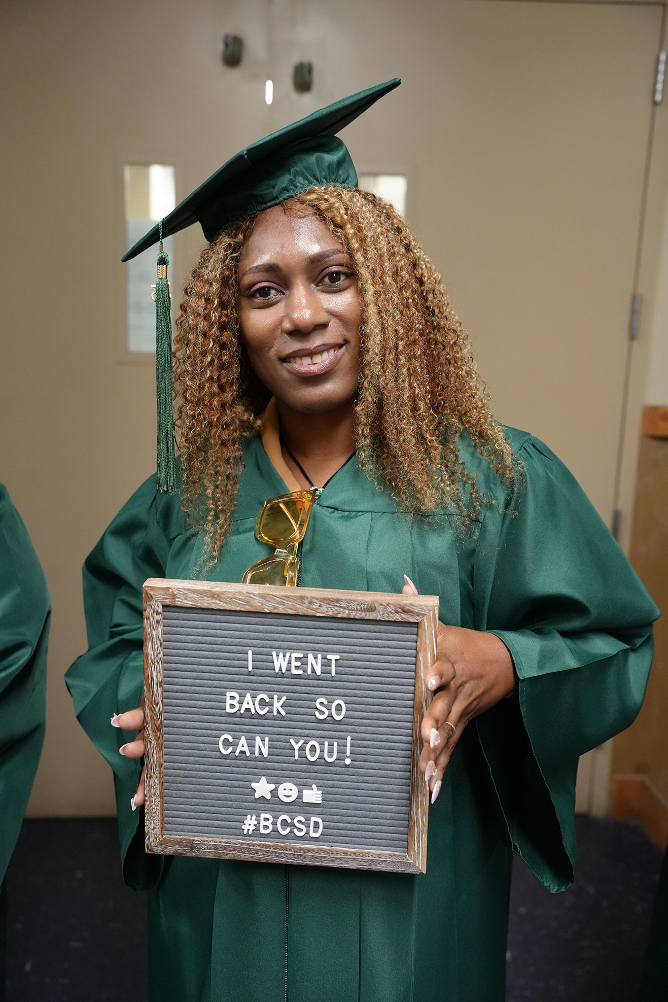 A woman in a green gown holds a sign that reads "I WENT BACK SO YOU CAN YOU!" A second person is partially visible.
