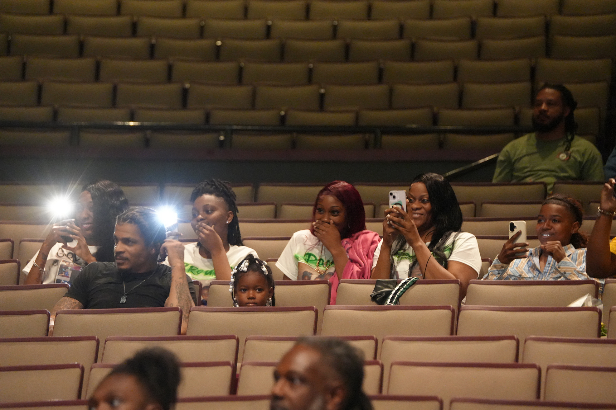 A group of people sits in theater seats, many holding up phones with flashlights. Empty seats surround them.