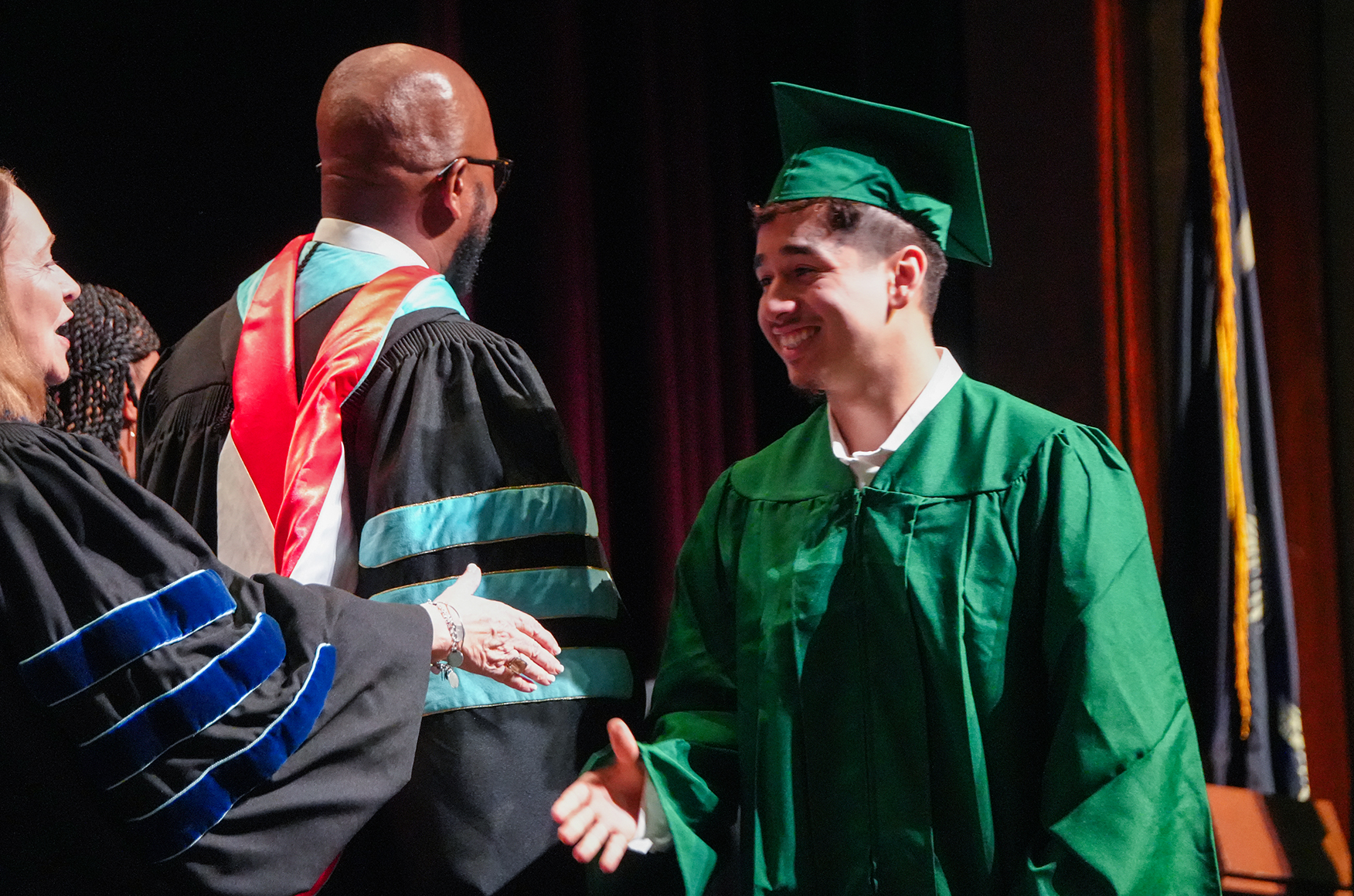 A man in a green graduation gown smiles at a woman in a black gown as a faculty member in red and blue gown shakes his hand.