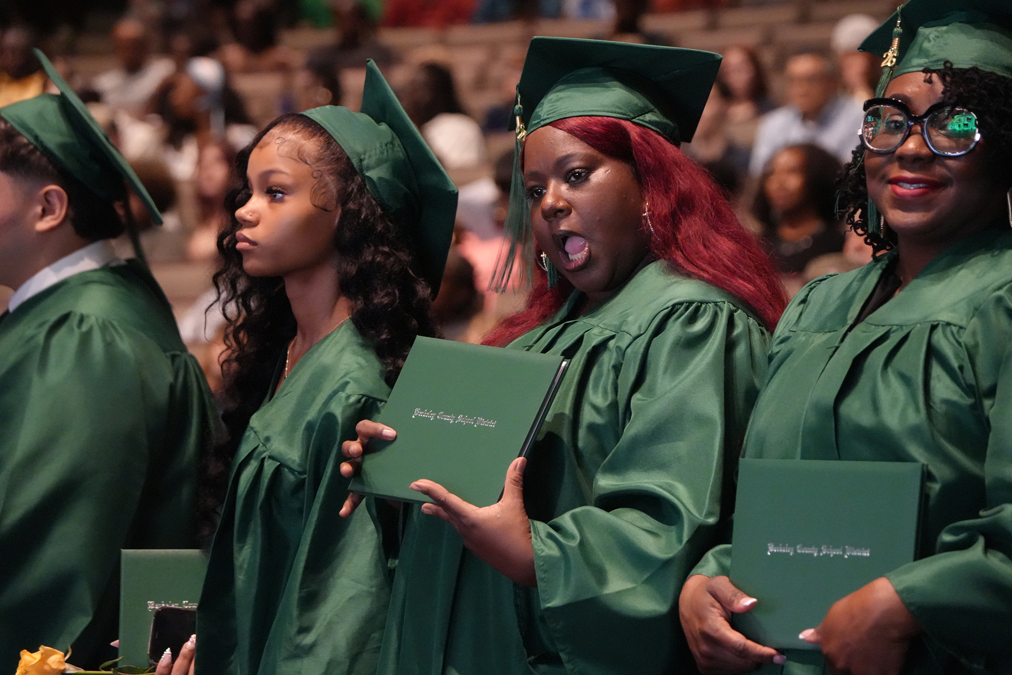 Four individuals in green graduation gowns and caps stand in a row. They hold green booklets. Behind them, people sit in bleachers, watching the ceremony.