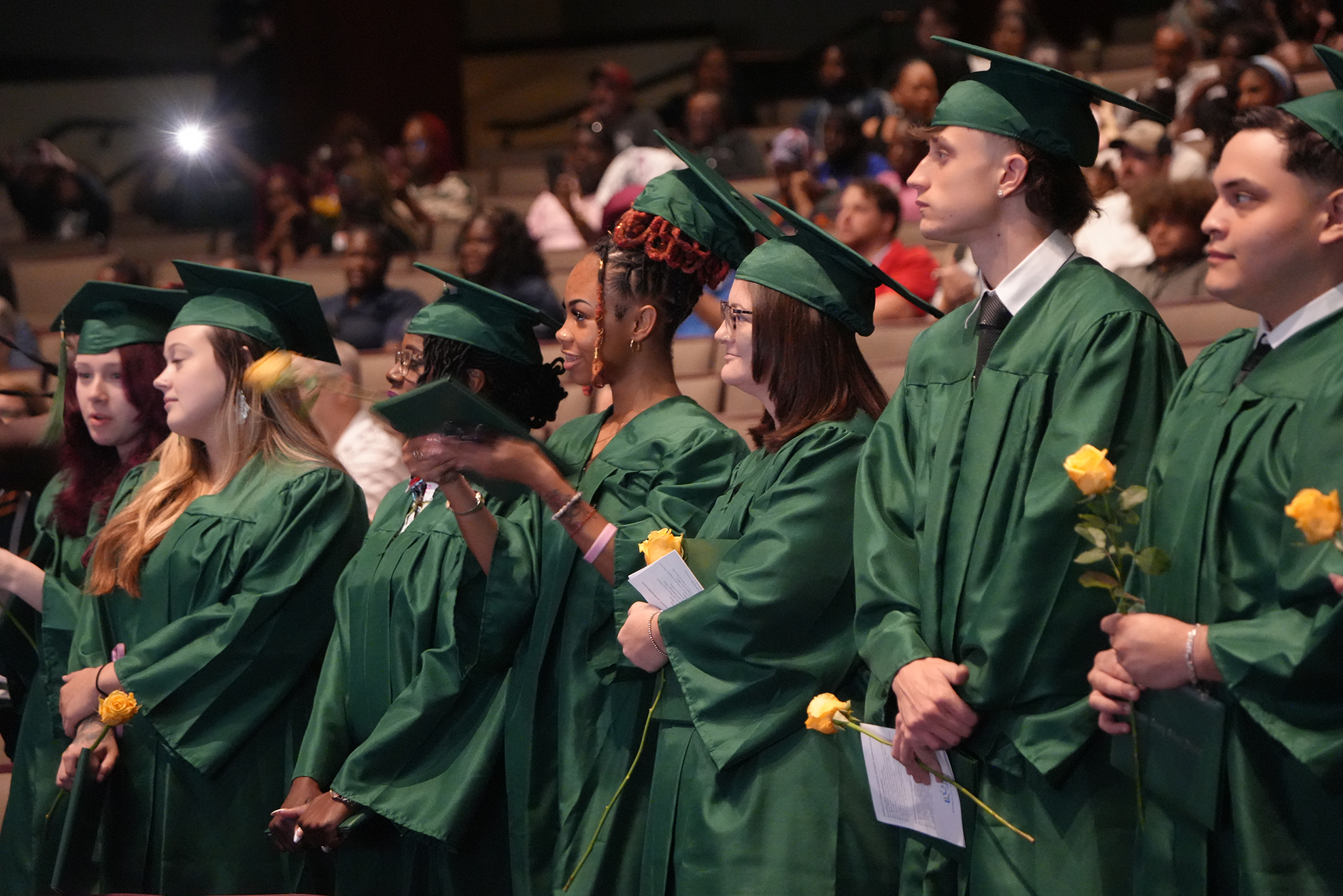 A line of students in green graduation gowns holding yellow flowers in an auditorium with a blurred audience.