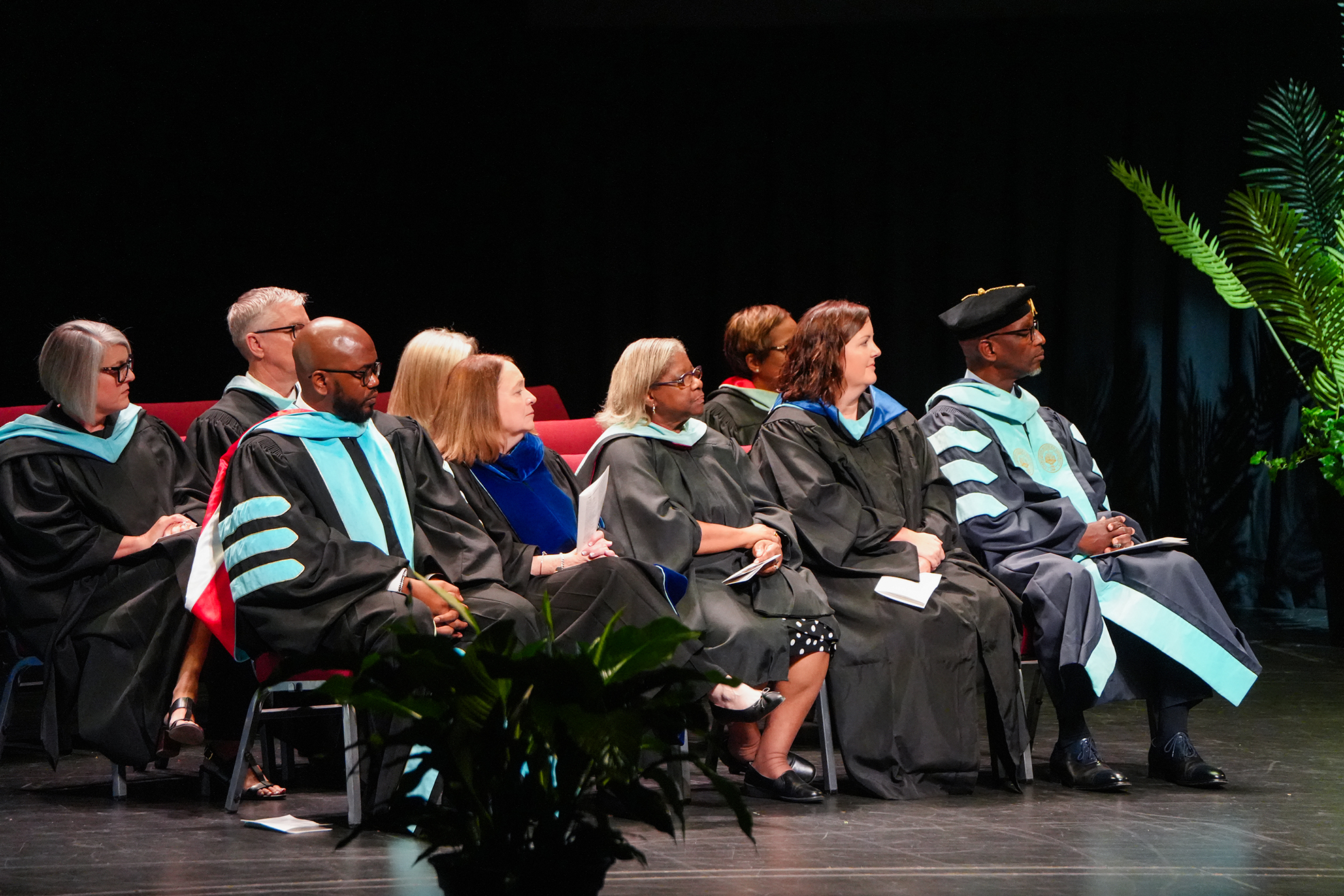 A group of people in academic regalia seated on a stage, possibly attending a graduation ceremony.