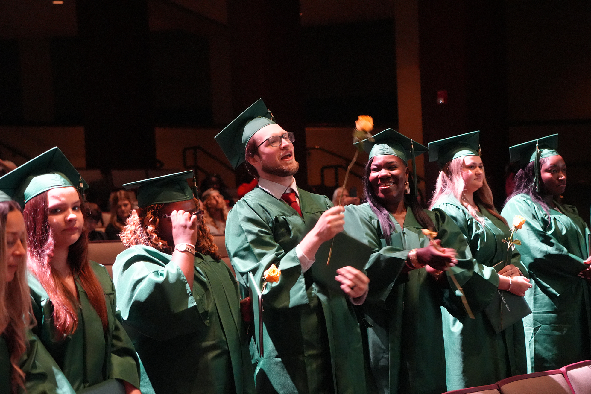 A group of graduates in green gowns and caps stand in a line at an indoor event.