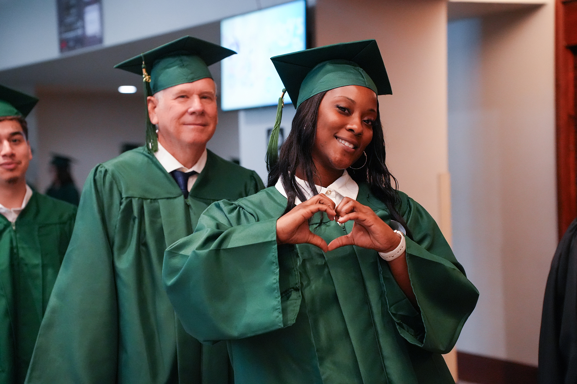 A woman and man in green graduation gowns and caps smile, with the woman making a heart shape with her hands.