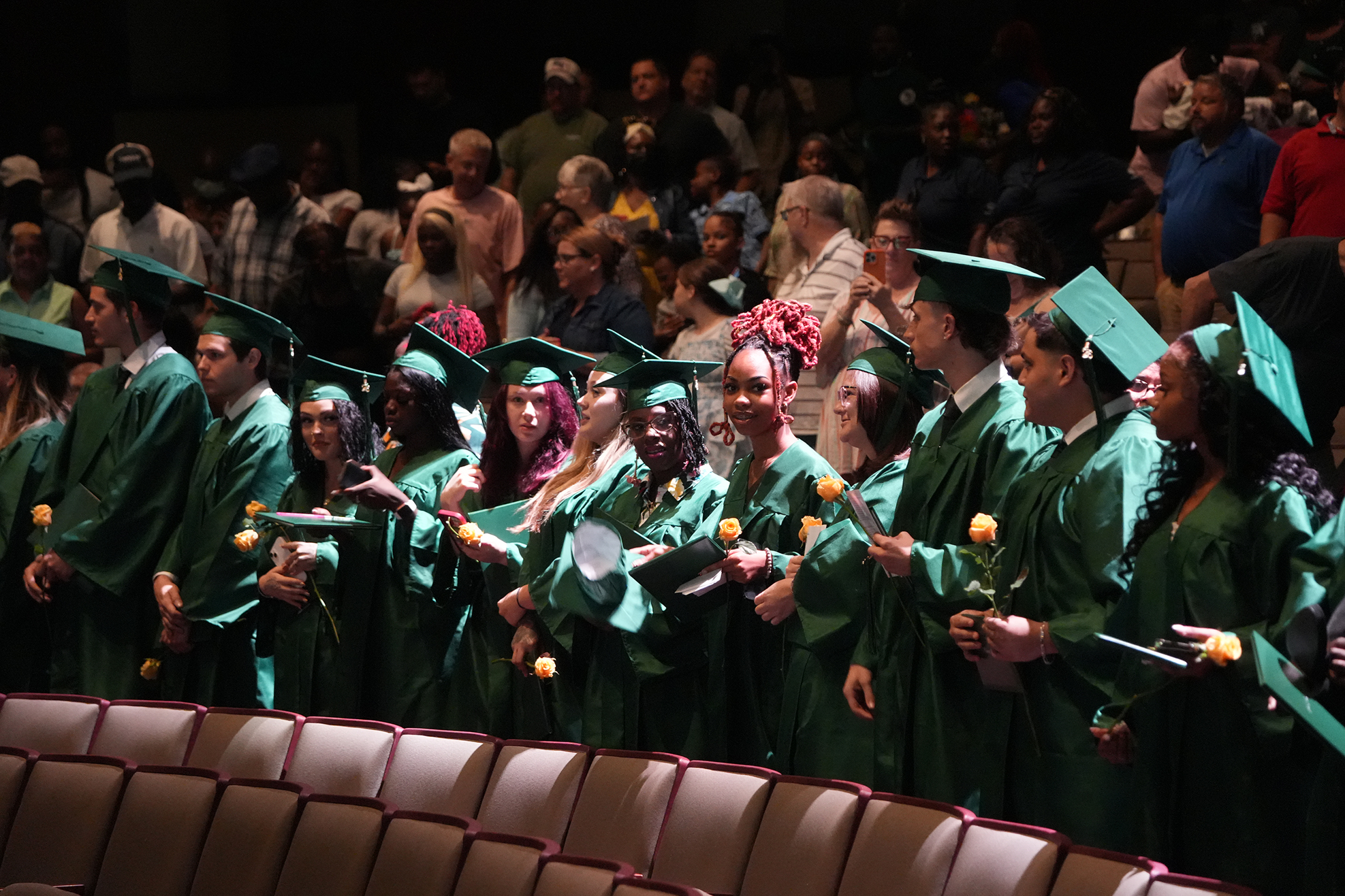 A group of people in green graduation gowns holding yellow flowers stands in front of a brown seat.