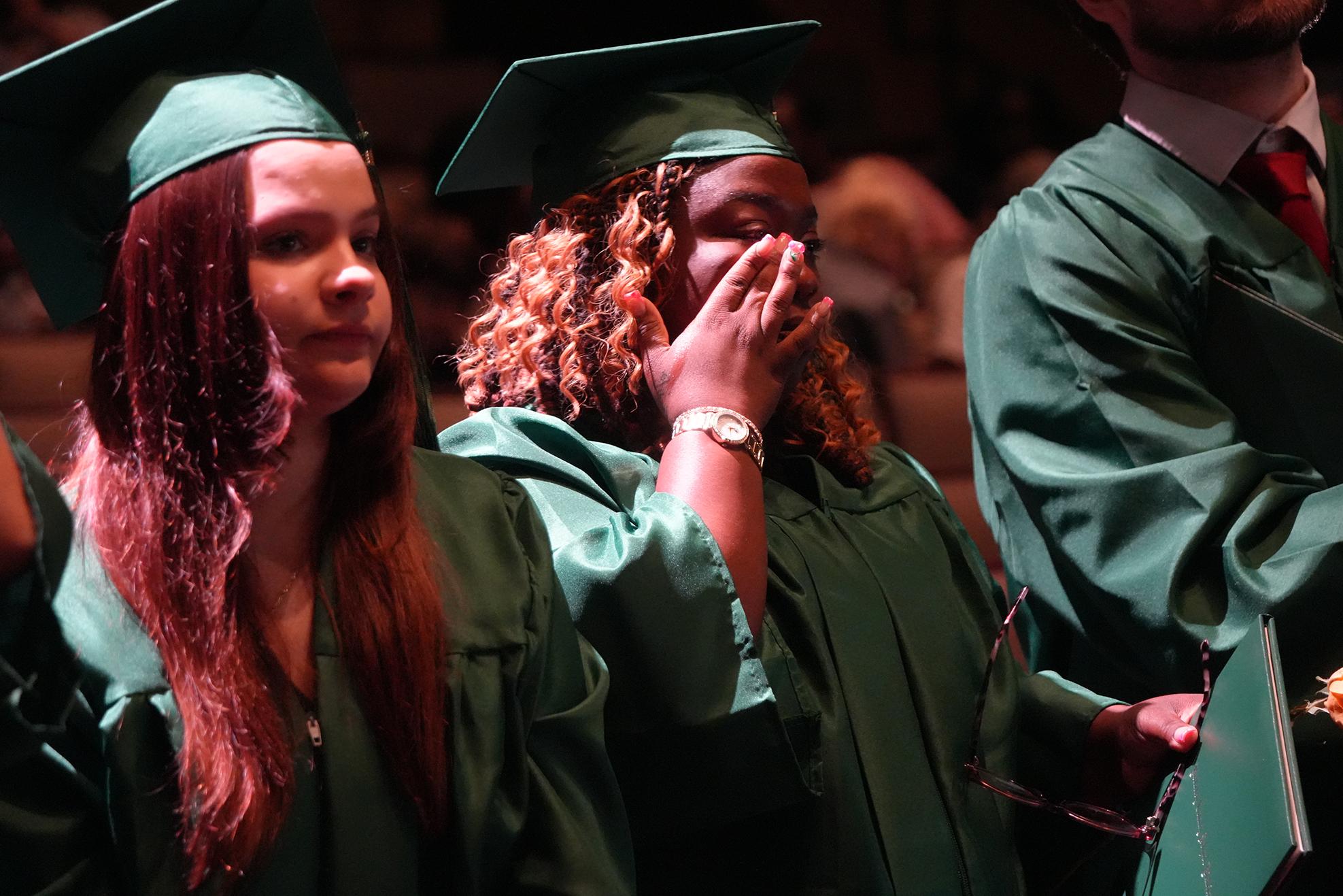 Graduates in green caps and gowns attend a ceremony; one covers mouth with hand.