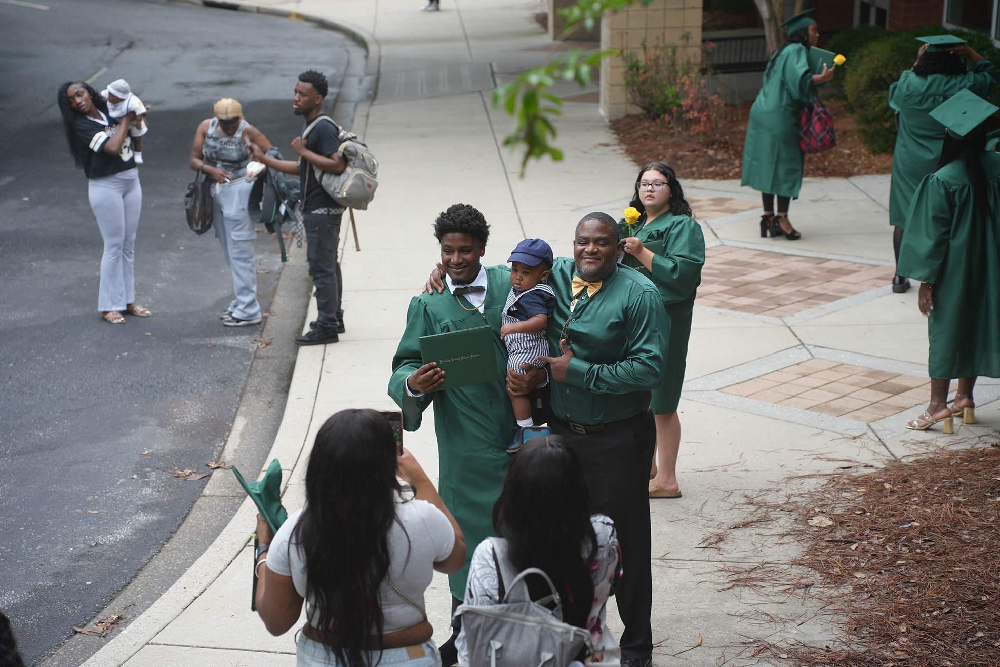 A group of people dressed in green graduation gowns and caps on a sidewalk. Some hold infants.