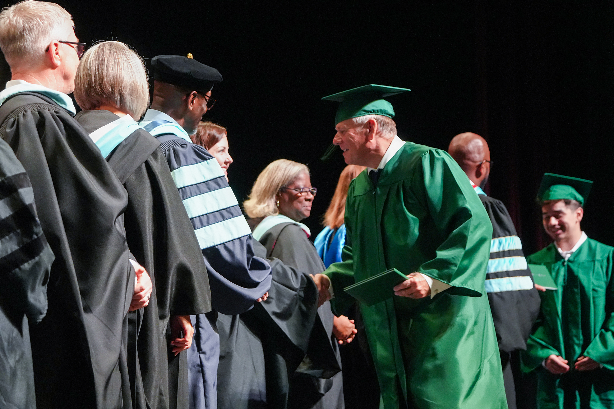 A group of people in graduation gowns and caps stand in line, one individual wearing a green gown shakes hands.