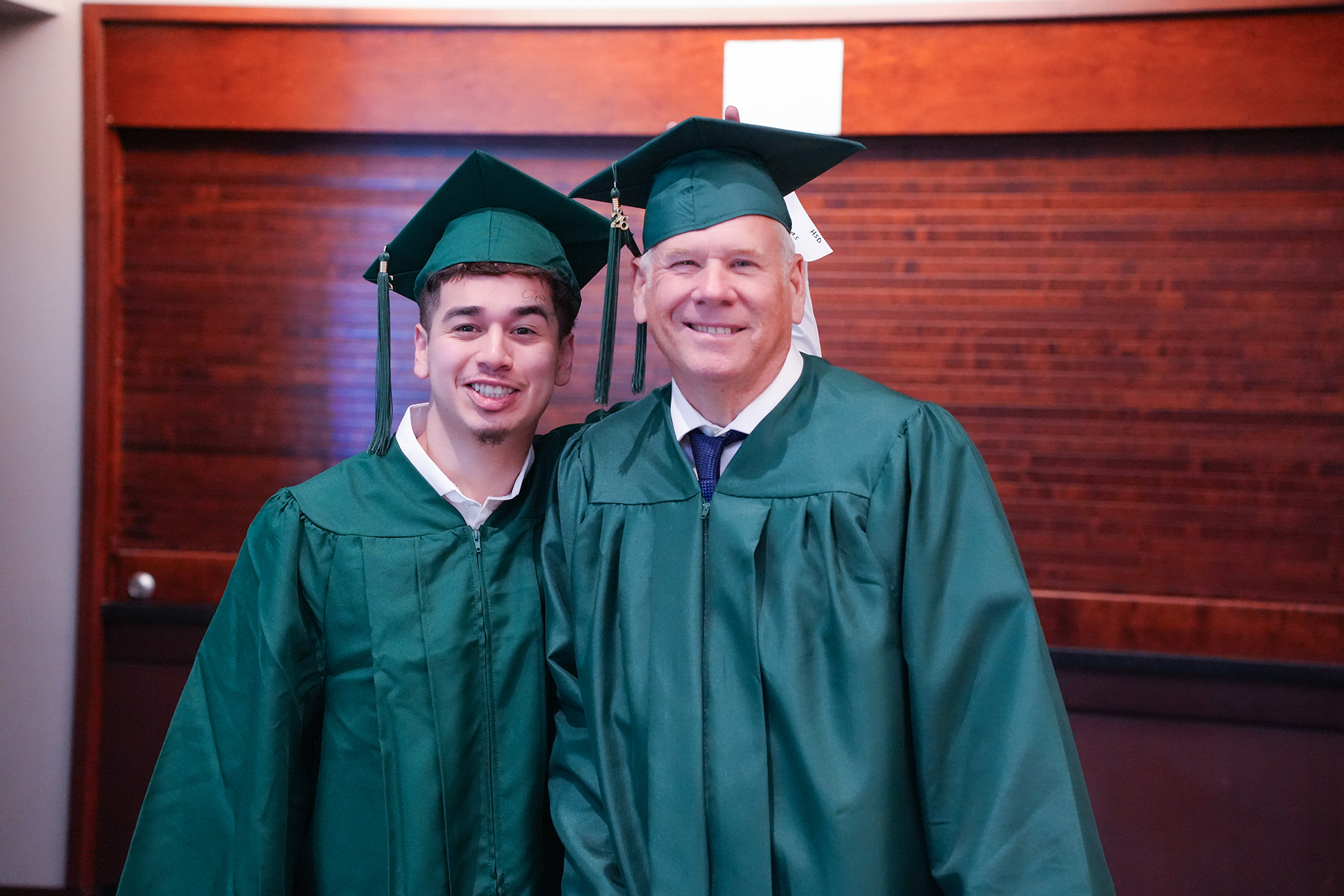 Two people in graduation gowns and caps smile for a photo. One person wears a tie. Background has wooden design.