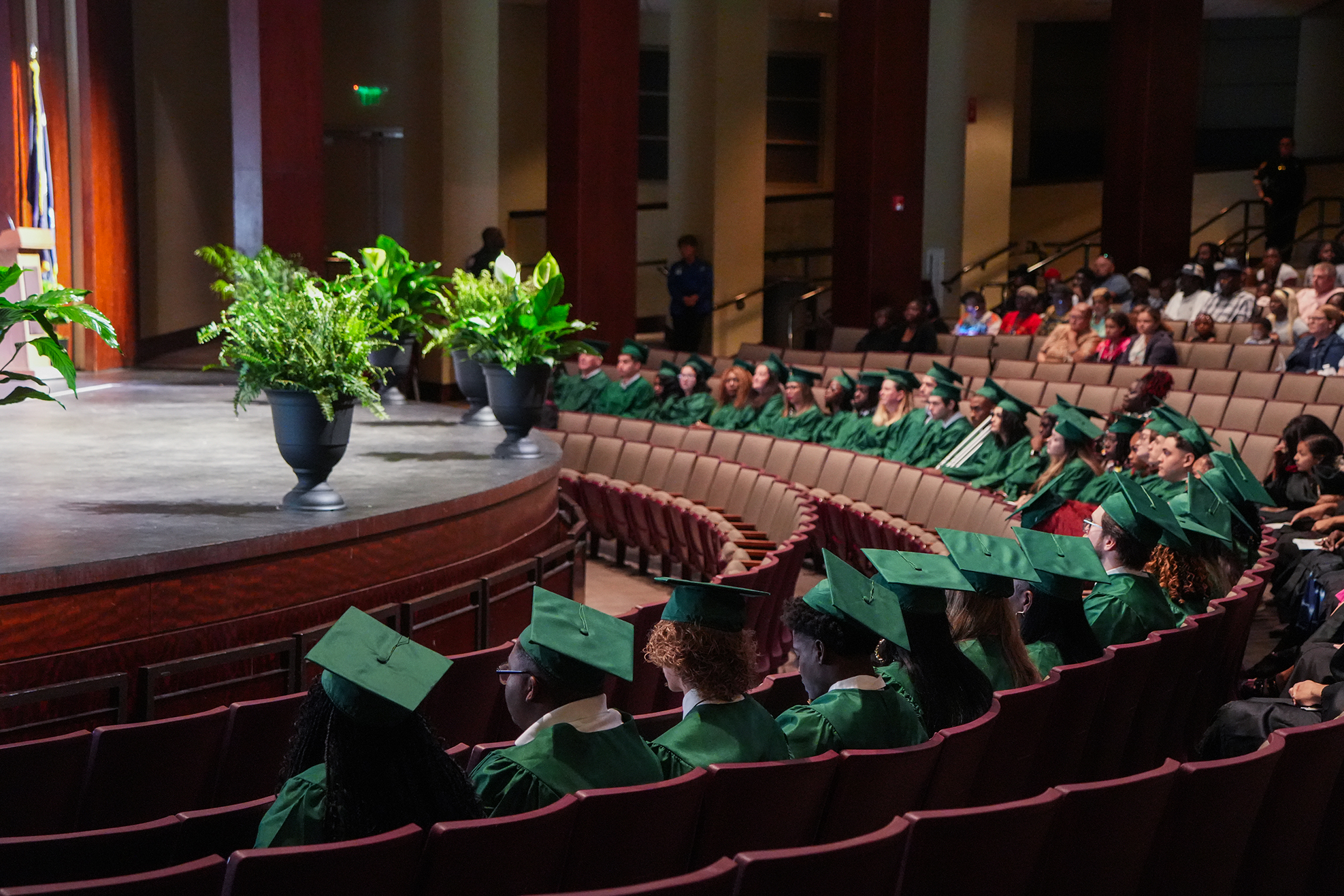 A group of people wearing green caps and gowns seated in rows, watching a graduation ceremony on stage.