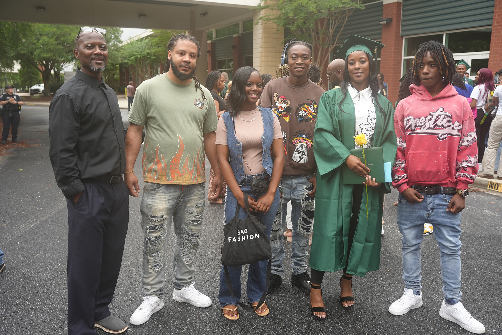 A group of people stand together in a parking lot, two wearing caps and gowns, one holding a flower.