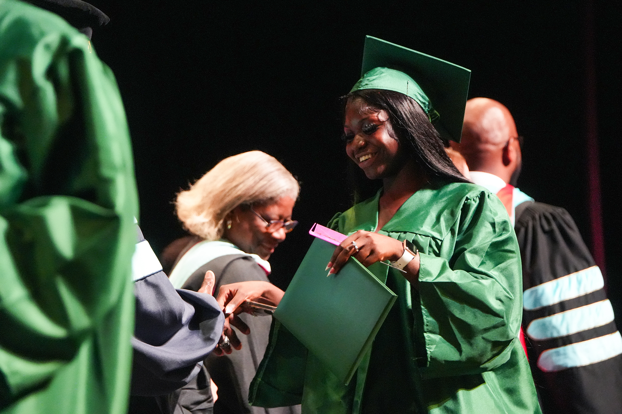 A woman in a green graduation gown holds a folder while standing among other people in green and black gowns.