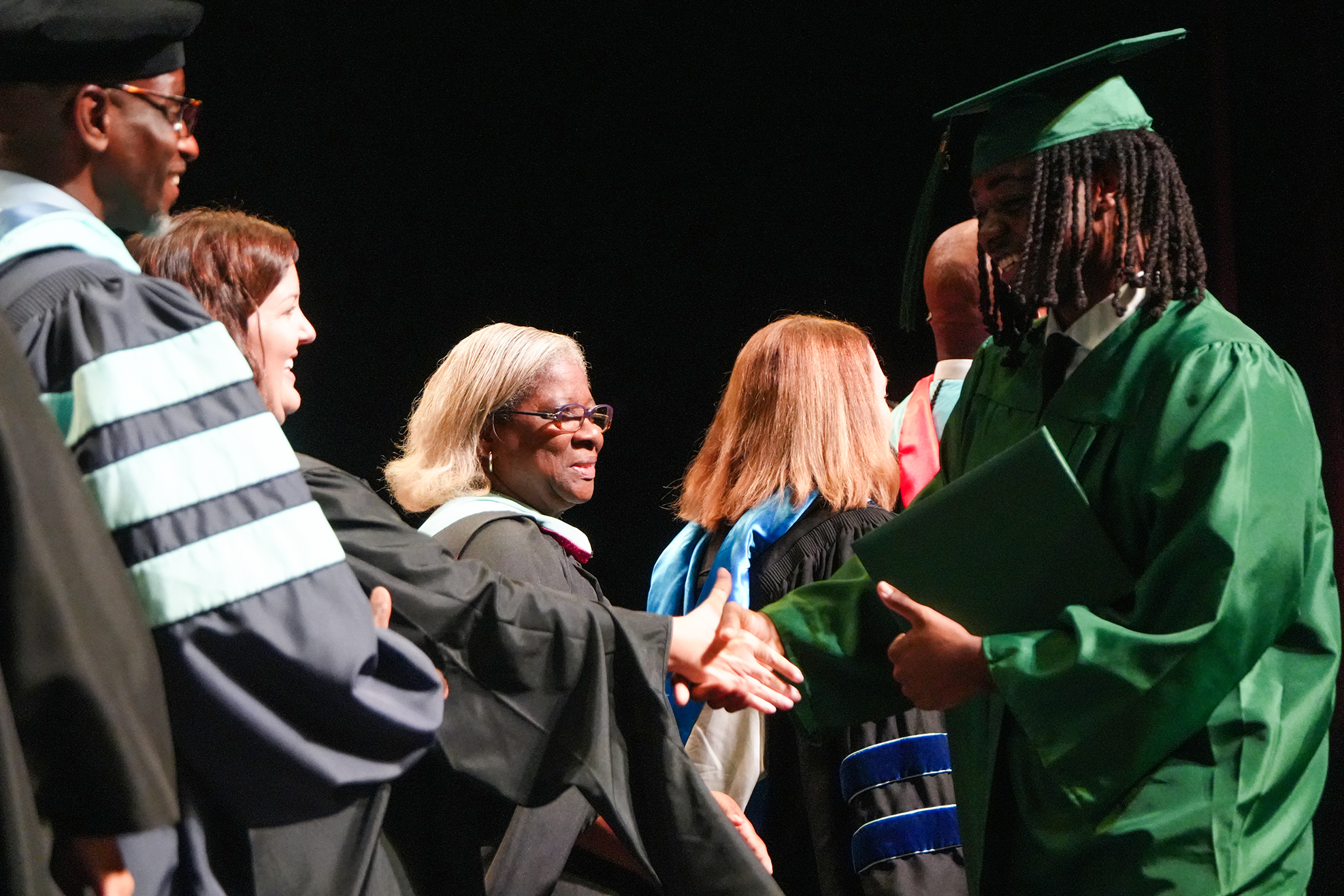 A graduate shakes hands with a faculty member, both wearing caps and gowns, while others observe.