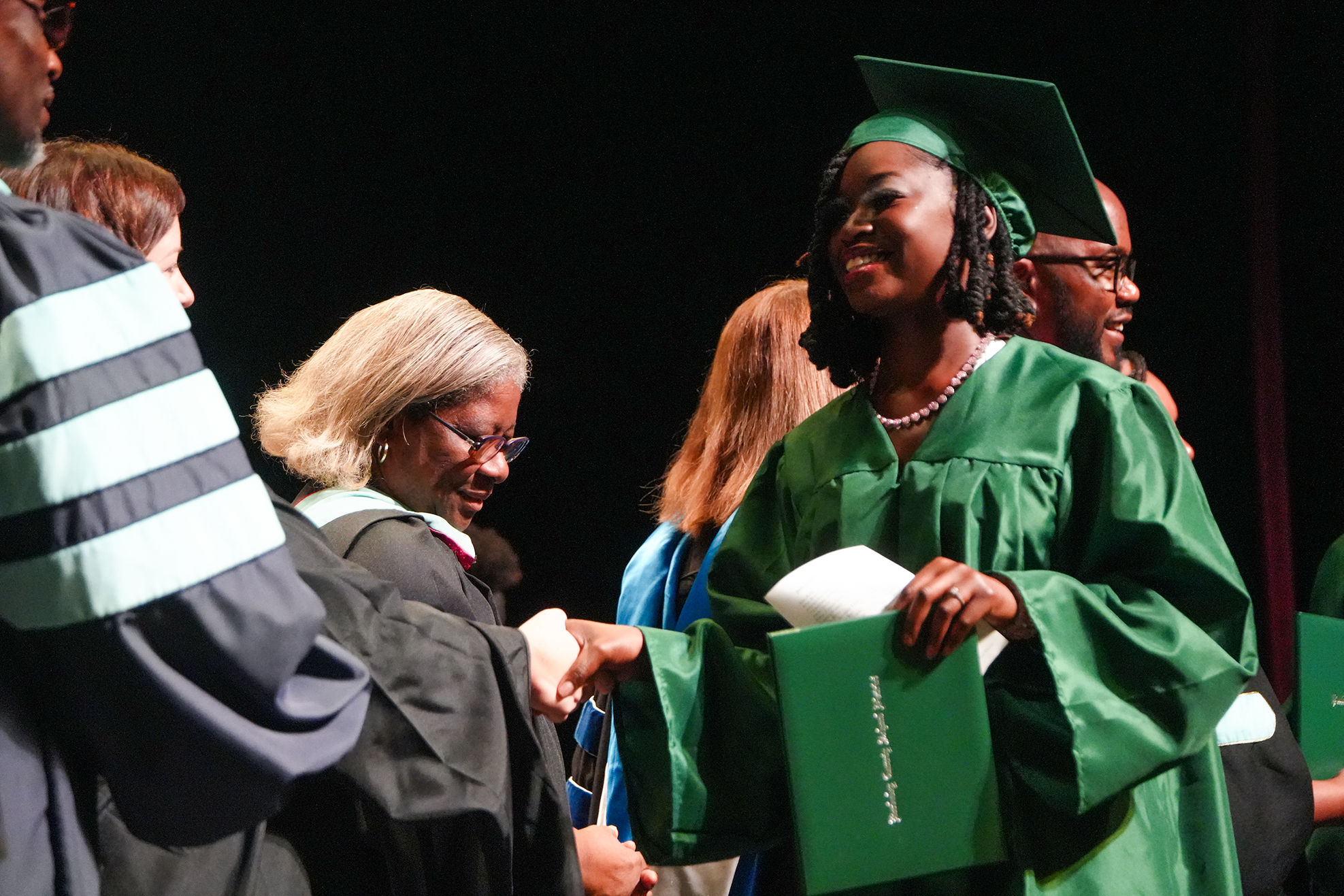A woman in a green graduation gown shakes hands with a faculty member at a ceremony.