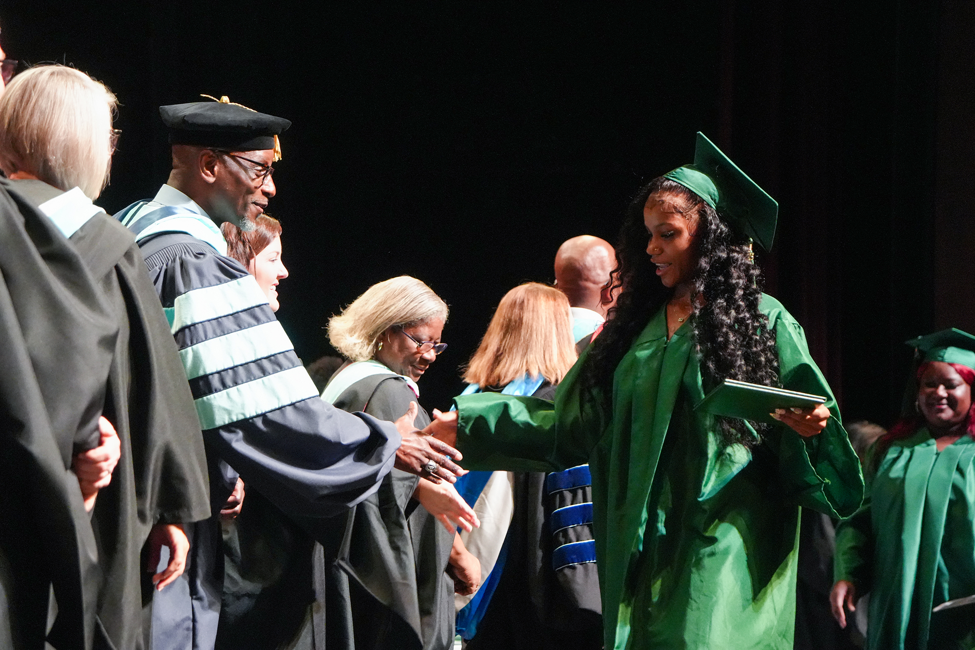 A woman in a green graduation gown is shaking hands with a man in a black and white gown.