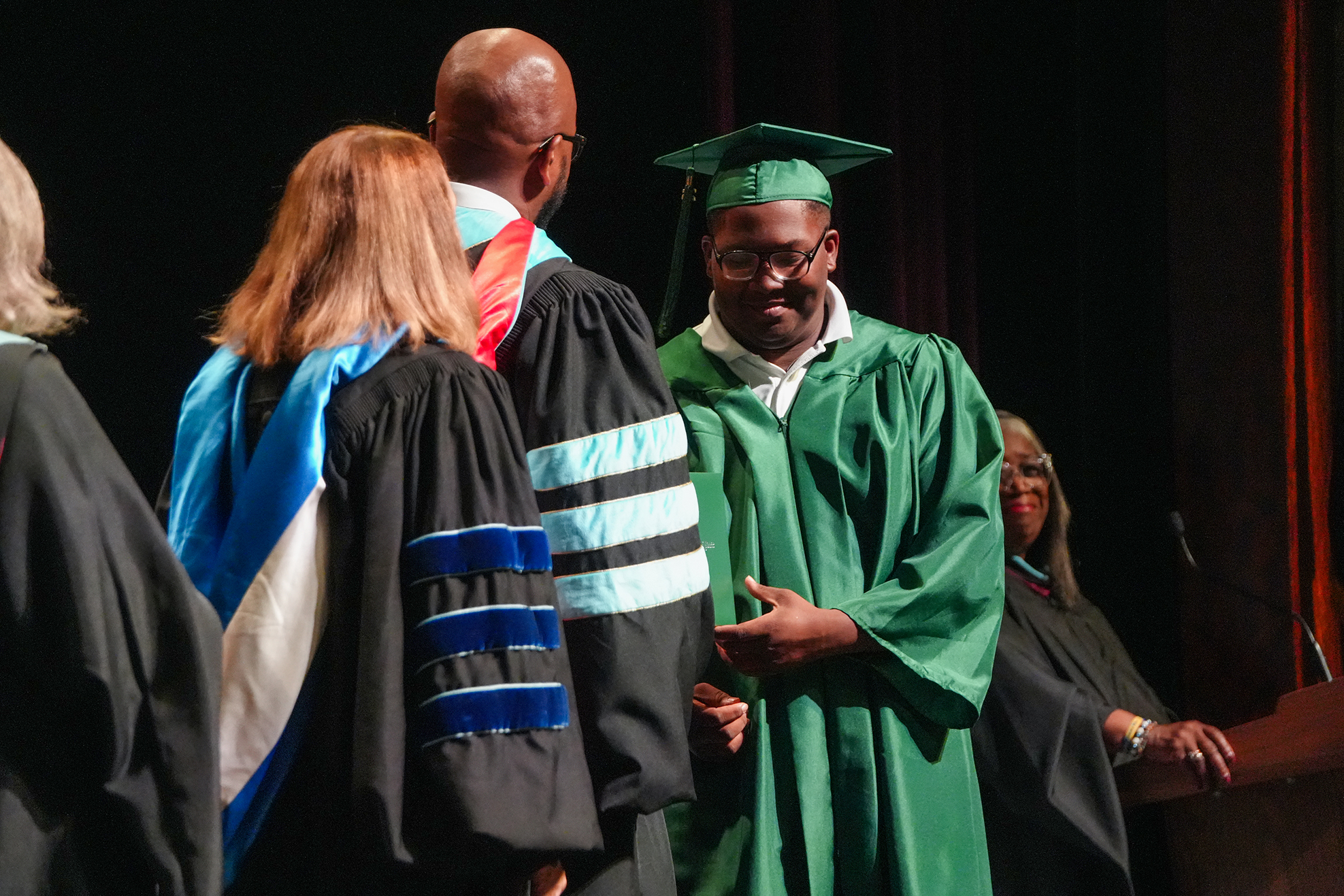 Several people in academic gowns, including a man in a green gown and cap, stand on a stage.