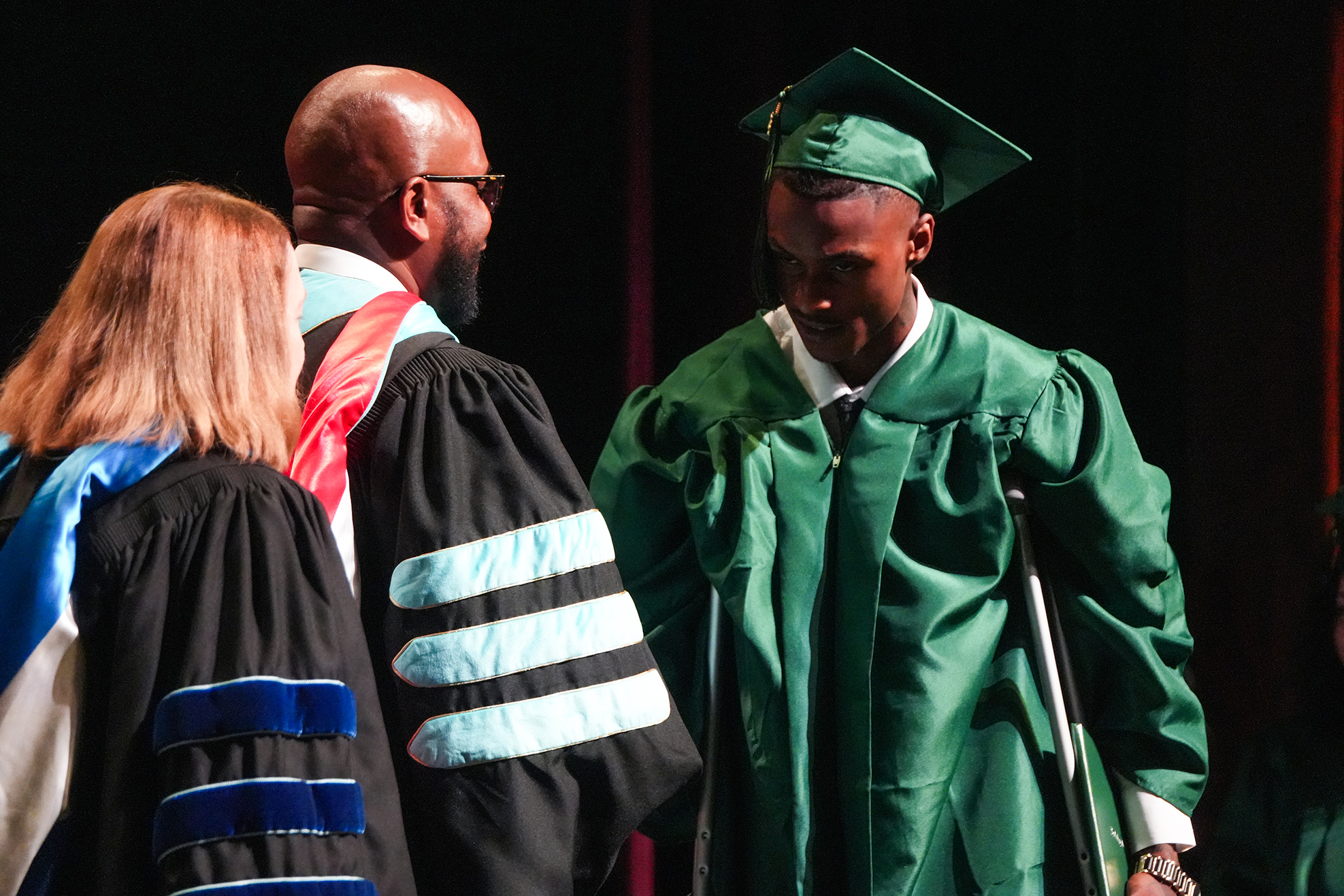 A man in a green graduation gown stands on stage using a crutch. He is surrounded by faculty members.