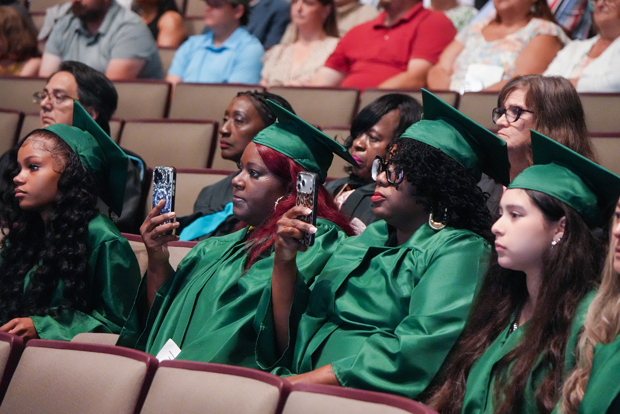 Several people sit in green graduation gowns and hats. They hold cellphones. Some are looking at screens.