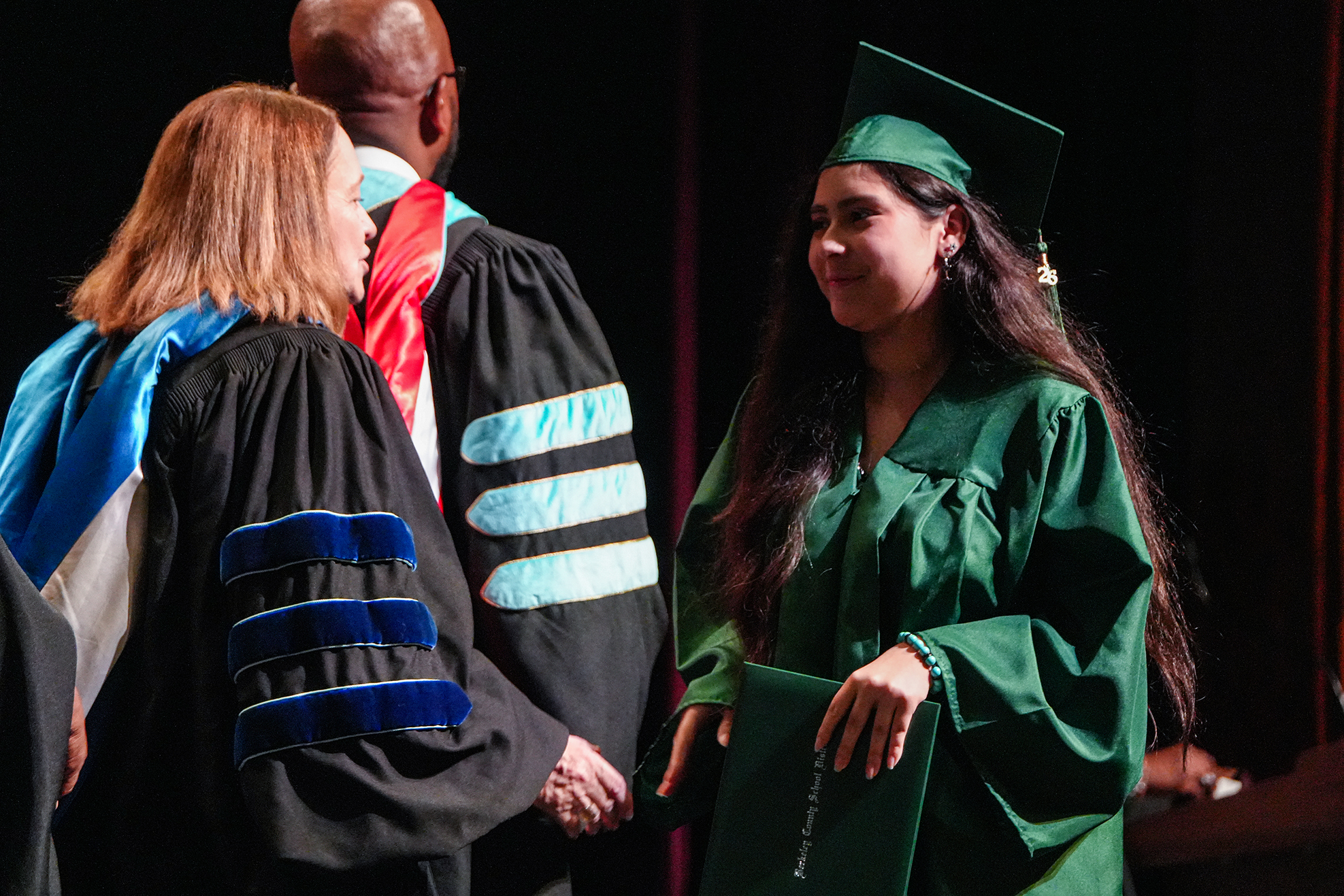 A woman in a green graduation gown smiles as she shakes hands with a person in a blue-trimmed black gown.