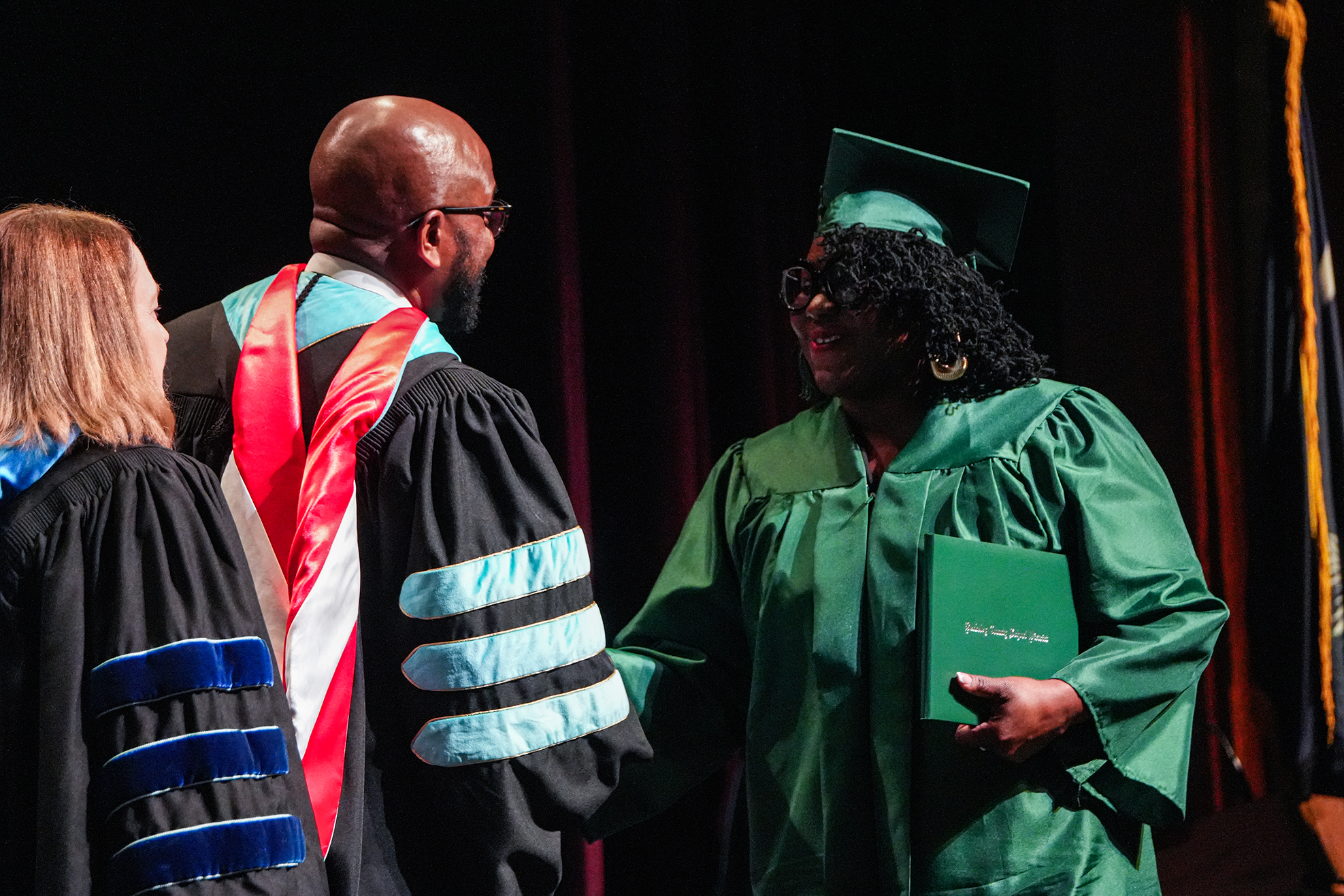A person in a green graduation gown stands with a book, smiling. Behind them, a man in a black and red gown stands.