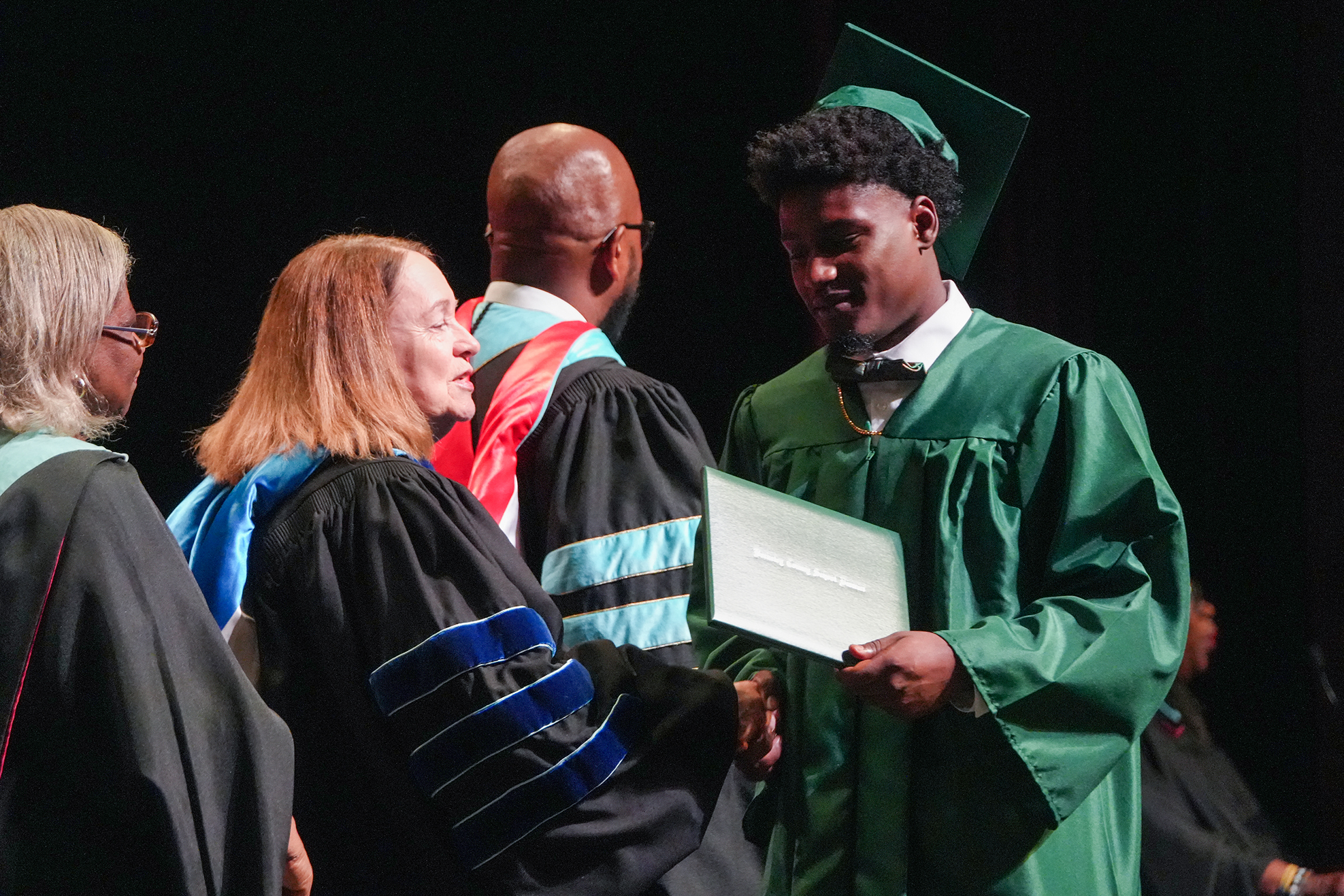 A man in a green gown receives a certificate from a woman in a blue gown. Other people in academic gowns stand nearby.