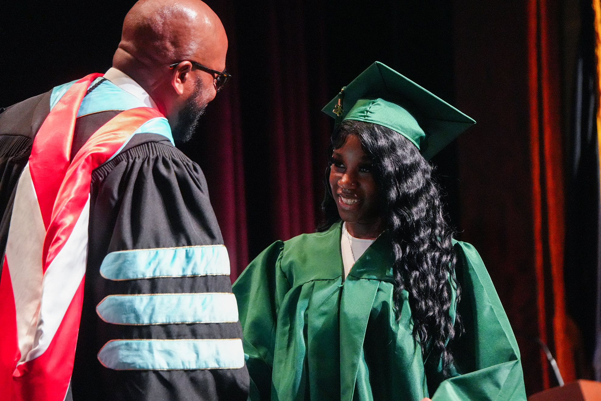 A graduate wearing a green gown and cap stands next to a faculty member in a black gown.