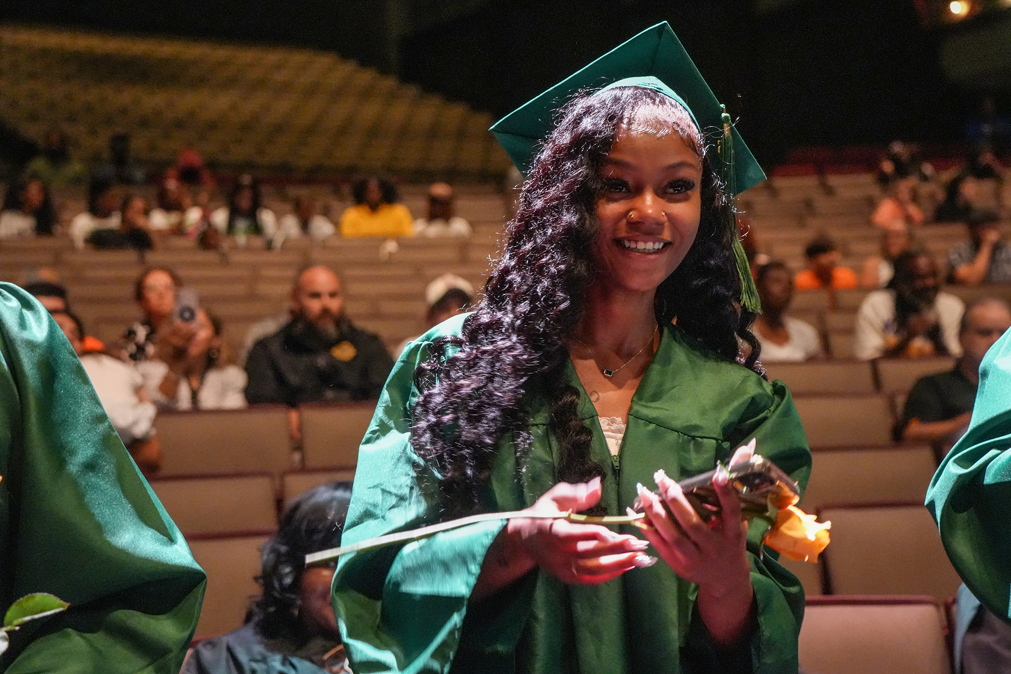 A woman in a green graduation gown holds flowers, smiling in a stadium with rows of spectators.