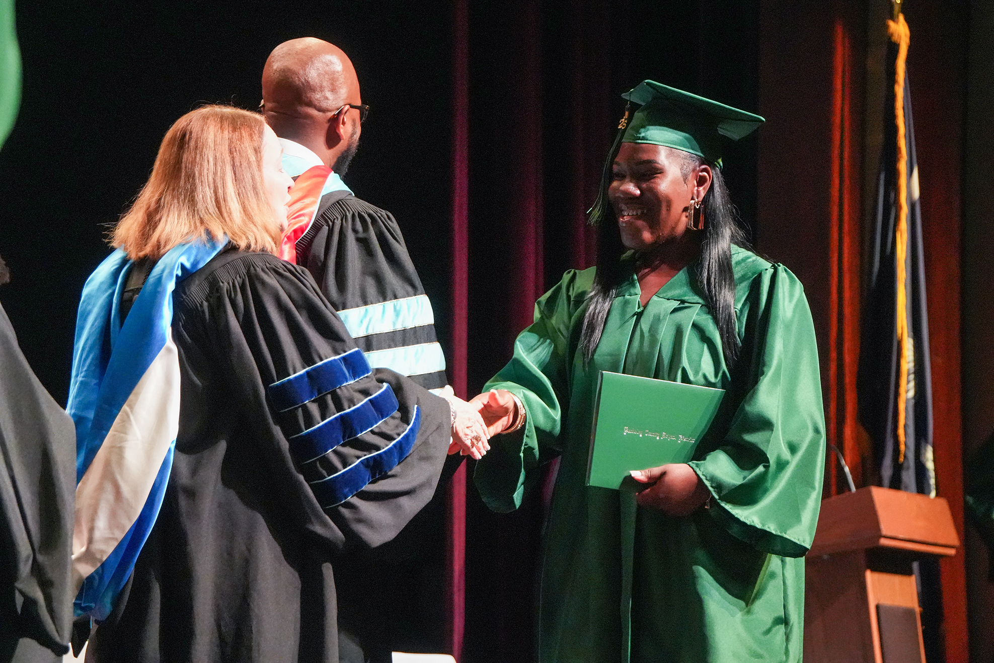 A person shakes hands with a graduate in a green gown, near a podium and flags.
