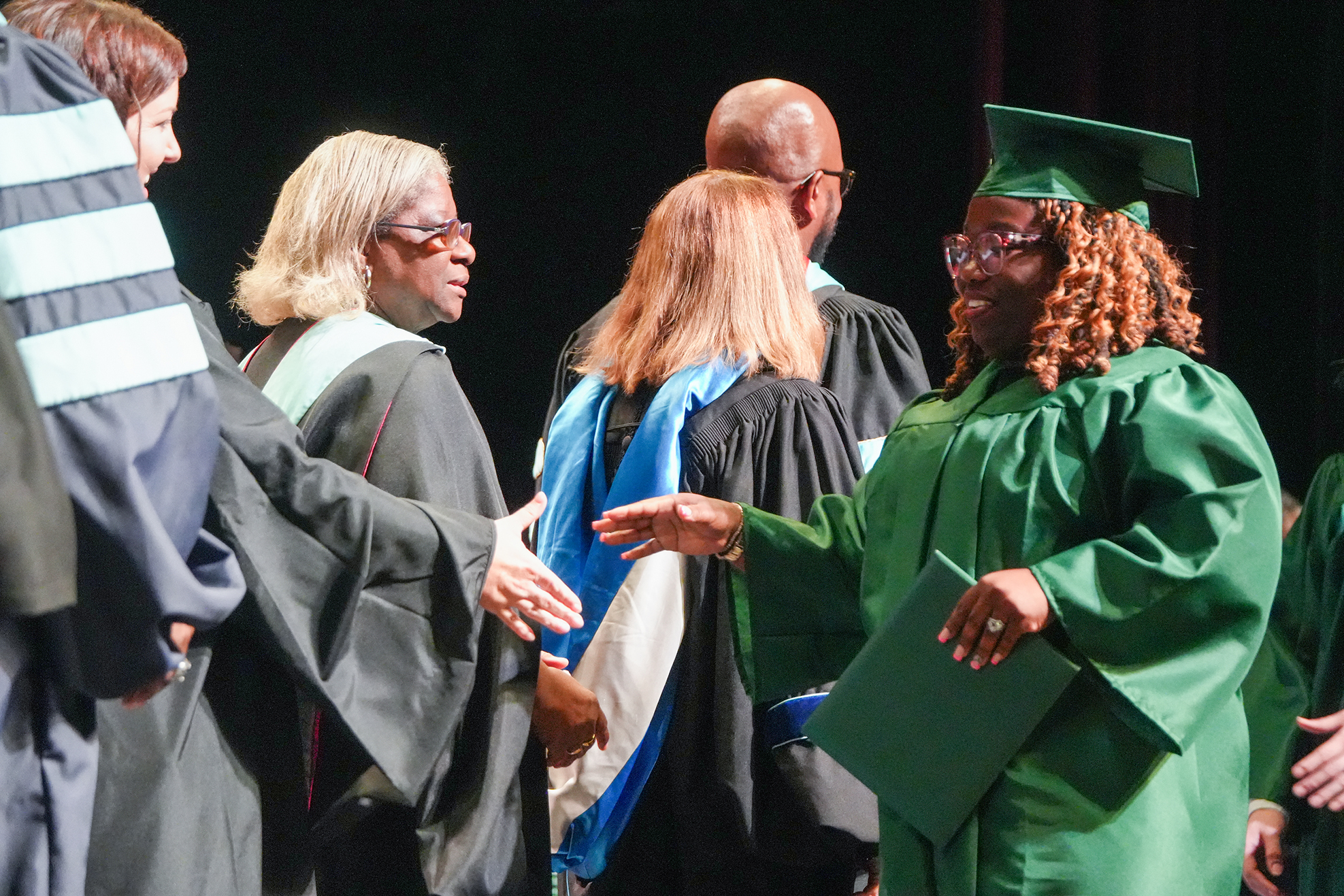 A graduate in a green gown congratulates others at a graduation ceremony. People wear black gowns and caps.